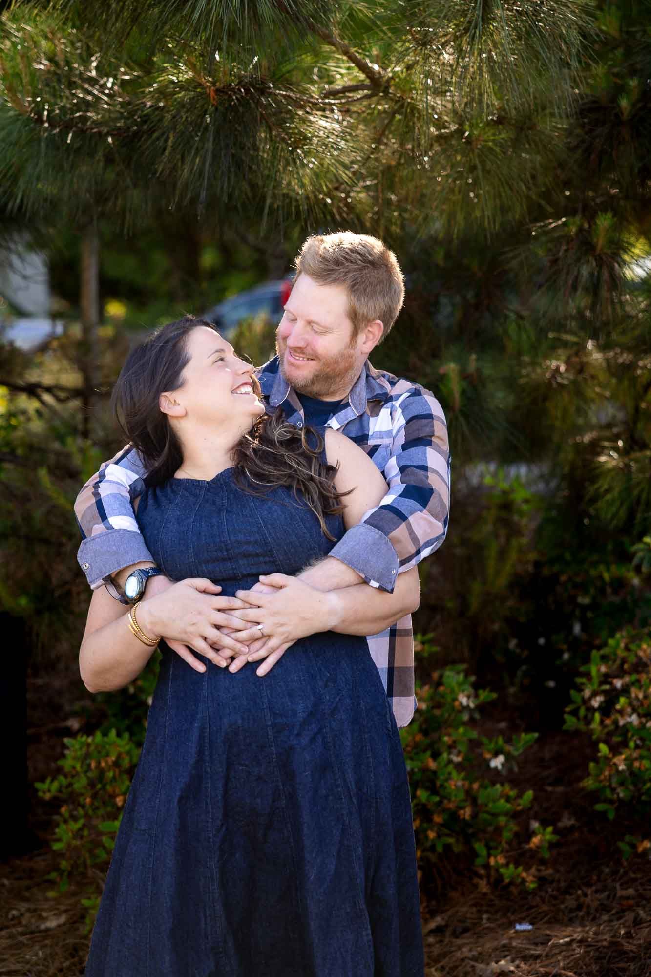 Happy couple embracing outdoors near pine trees, pregnant woman in navy dress smiling up at bearded man in plaid shirt.