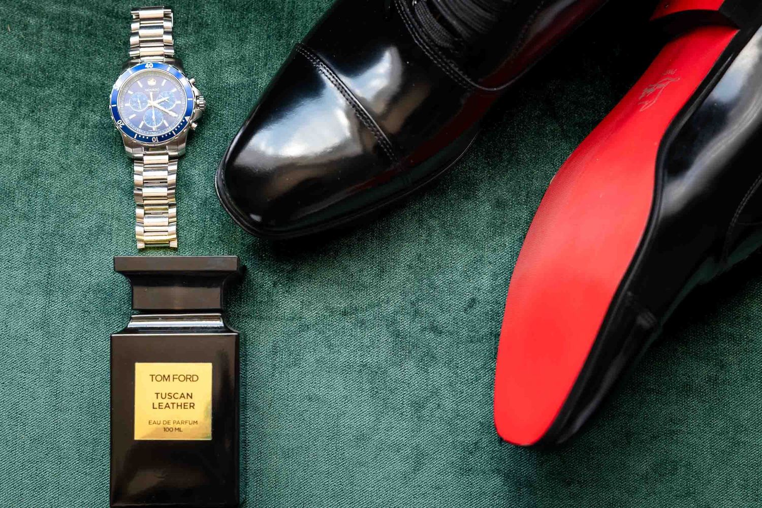 Tom Ford Tuscan Leather cologne beside a blue TAG Heuer watch and black Louboutin dress shoes on green velvet.