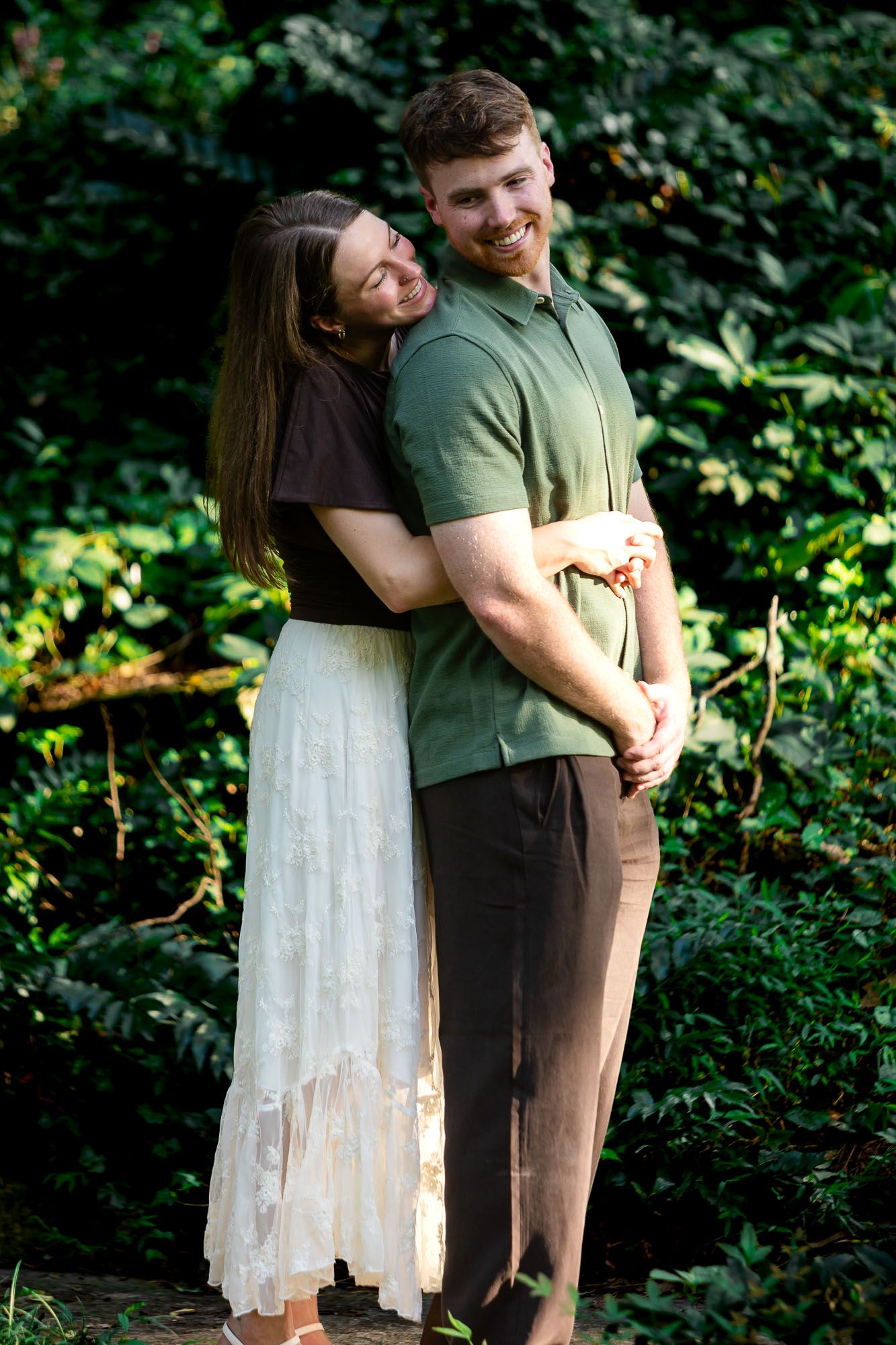 A smiling couple embraces outdoors, woman in white skirt hugging man in green polo, surrounded by lush green foliage.