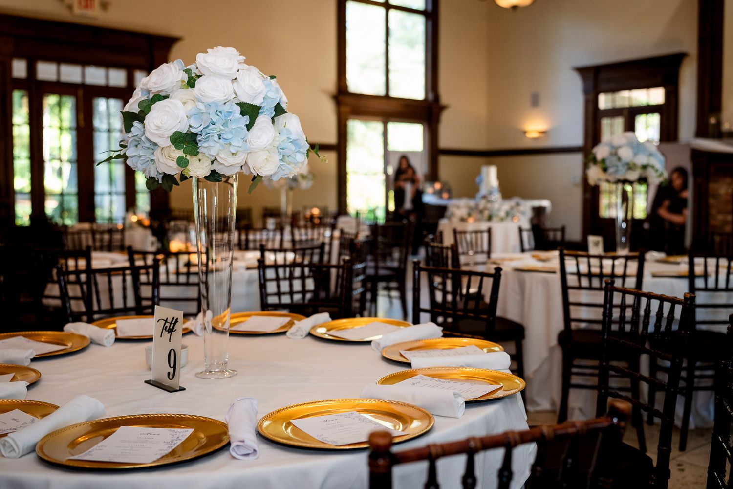 Elegant wedding reception hall with round tables, gold charger plates, white floral centerpieces, and dark Chiavari chairs.