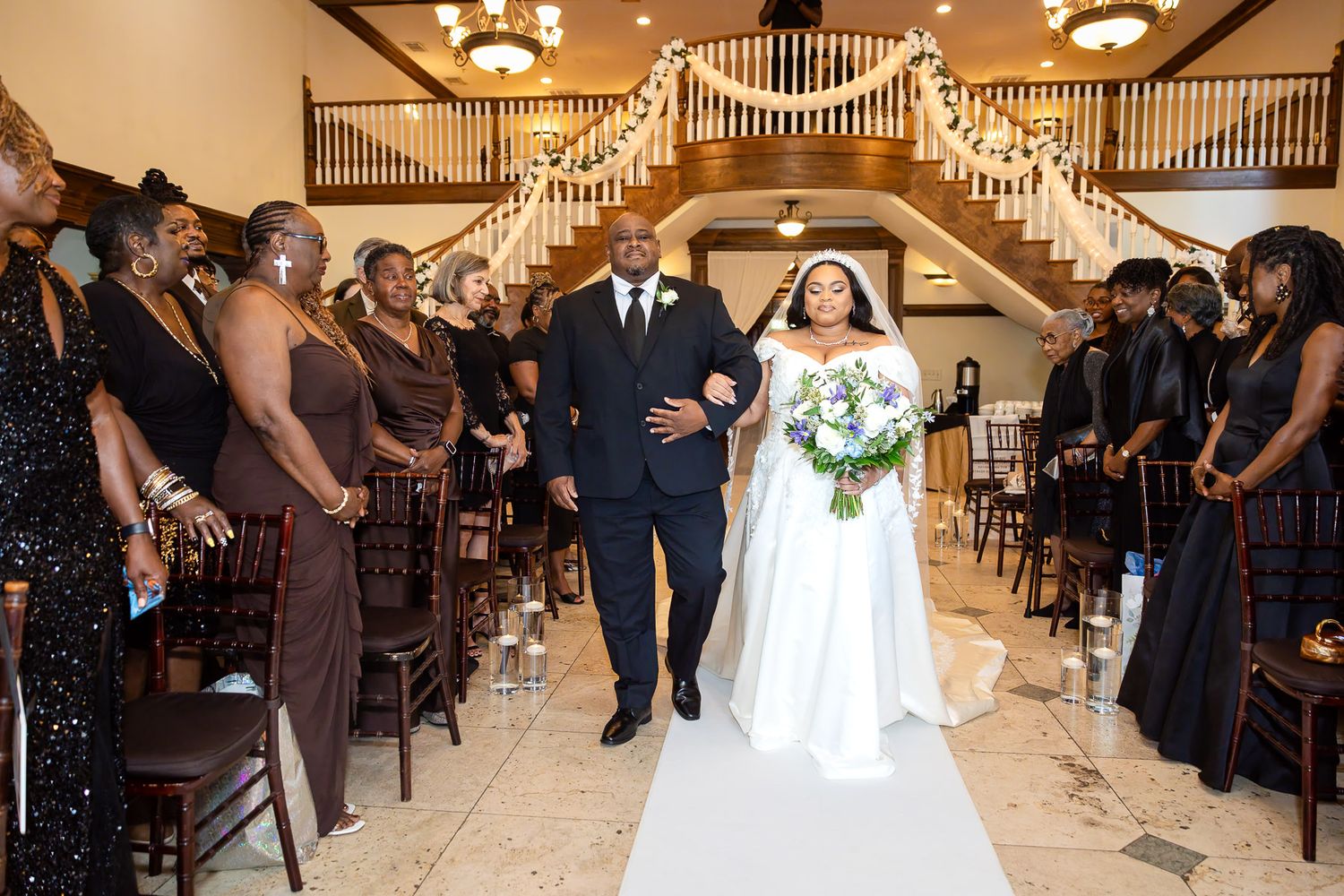 Bride and groom walk down the aisle at elegant wedding venue with grand staircase and guests in brown attire.