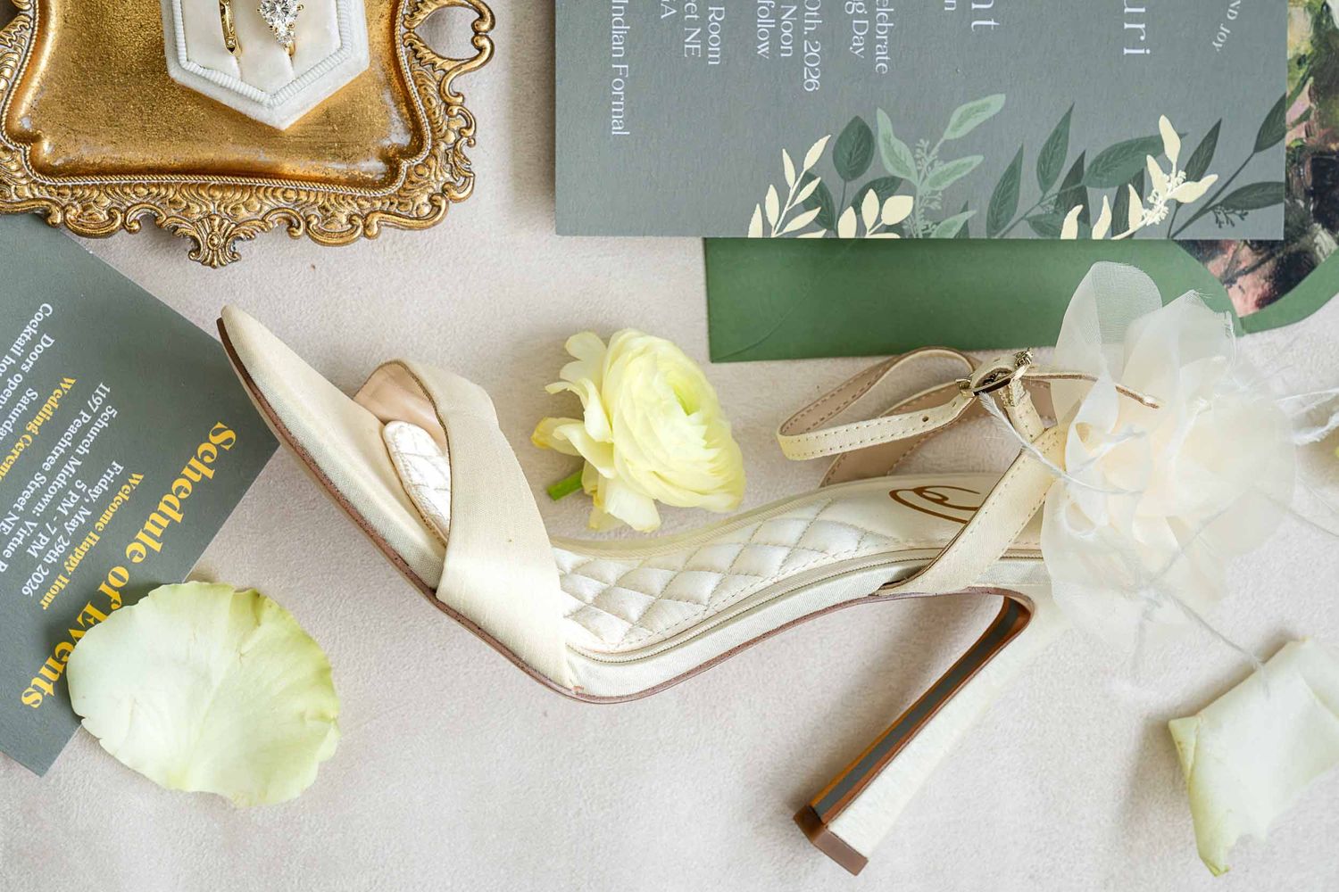 Elegant bridal flat lay with ivory quilted heels, green wedding invitations, white ranunculus, and gold ring dish.