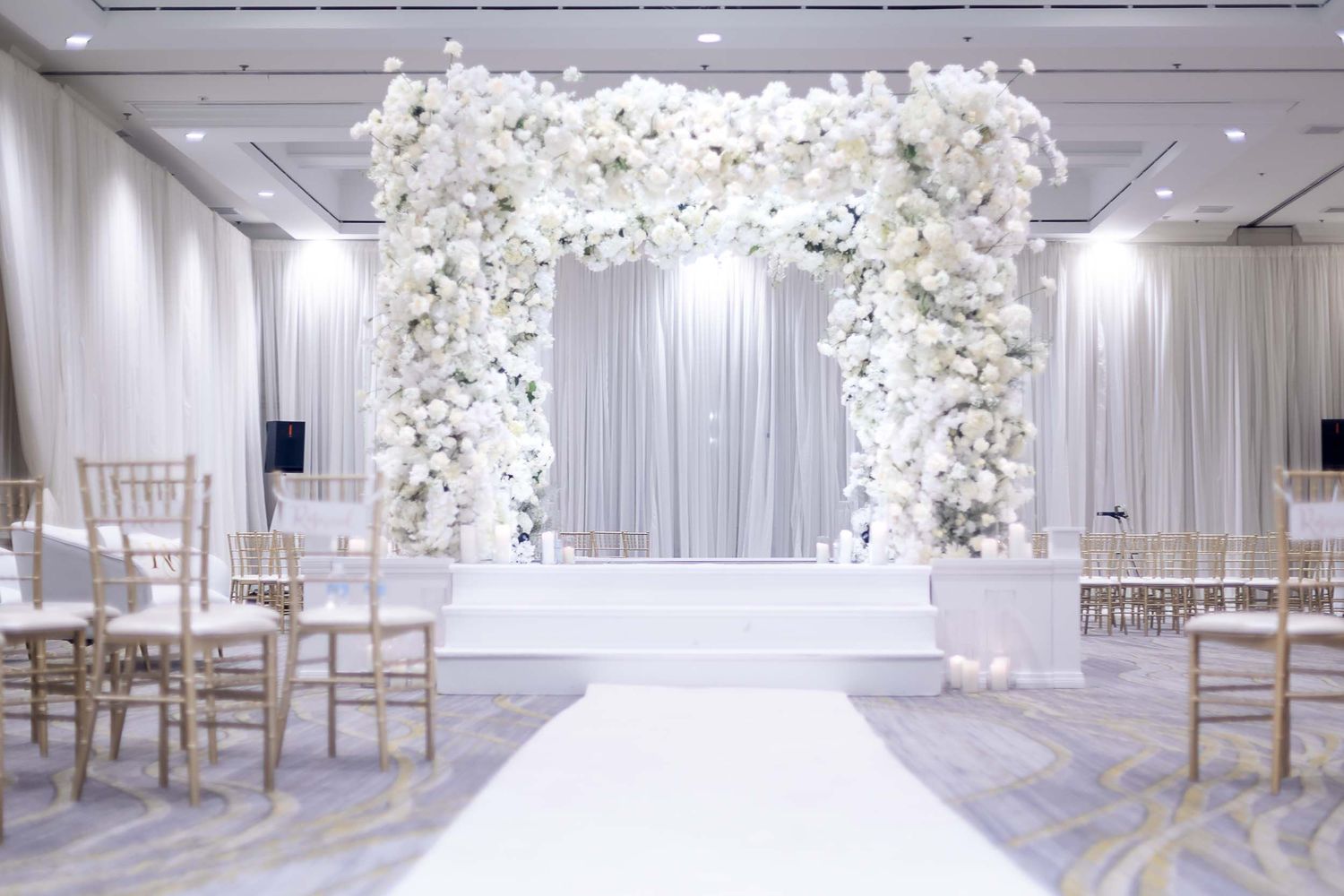 Elegant white wedding ceremony setup with floral arch, white aisle runner, and gold Chiavari chairs in a ballroom.