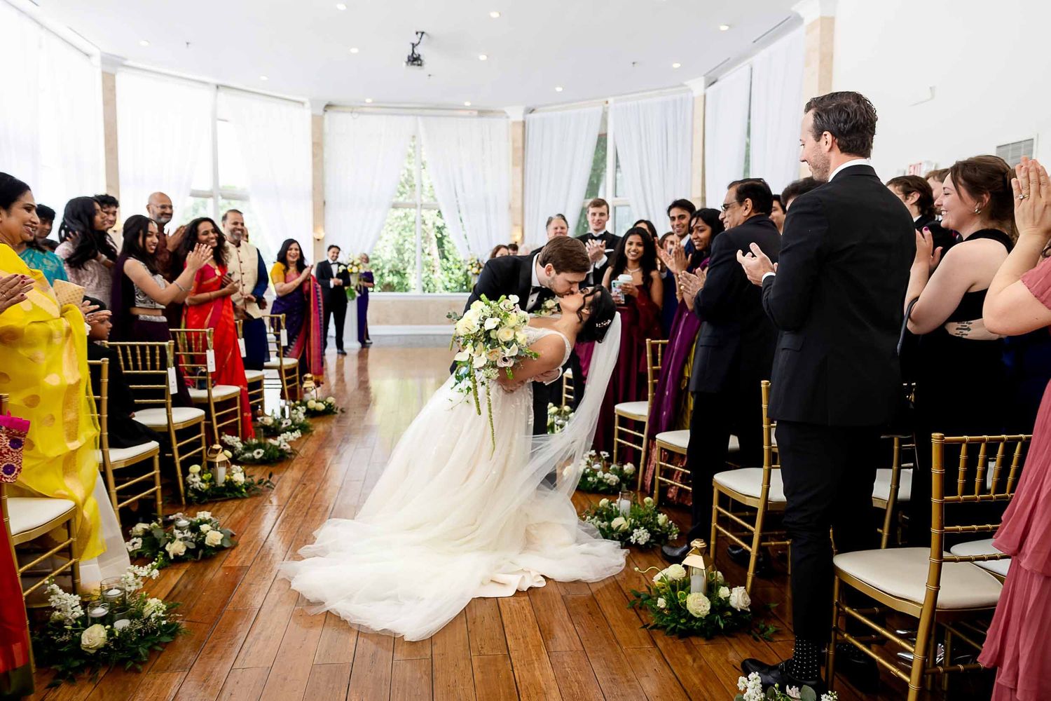 Bride and groom share first kiss at elegant indoor wedding ceremony with guests applauding in decorated aisle.