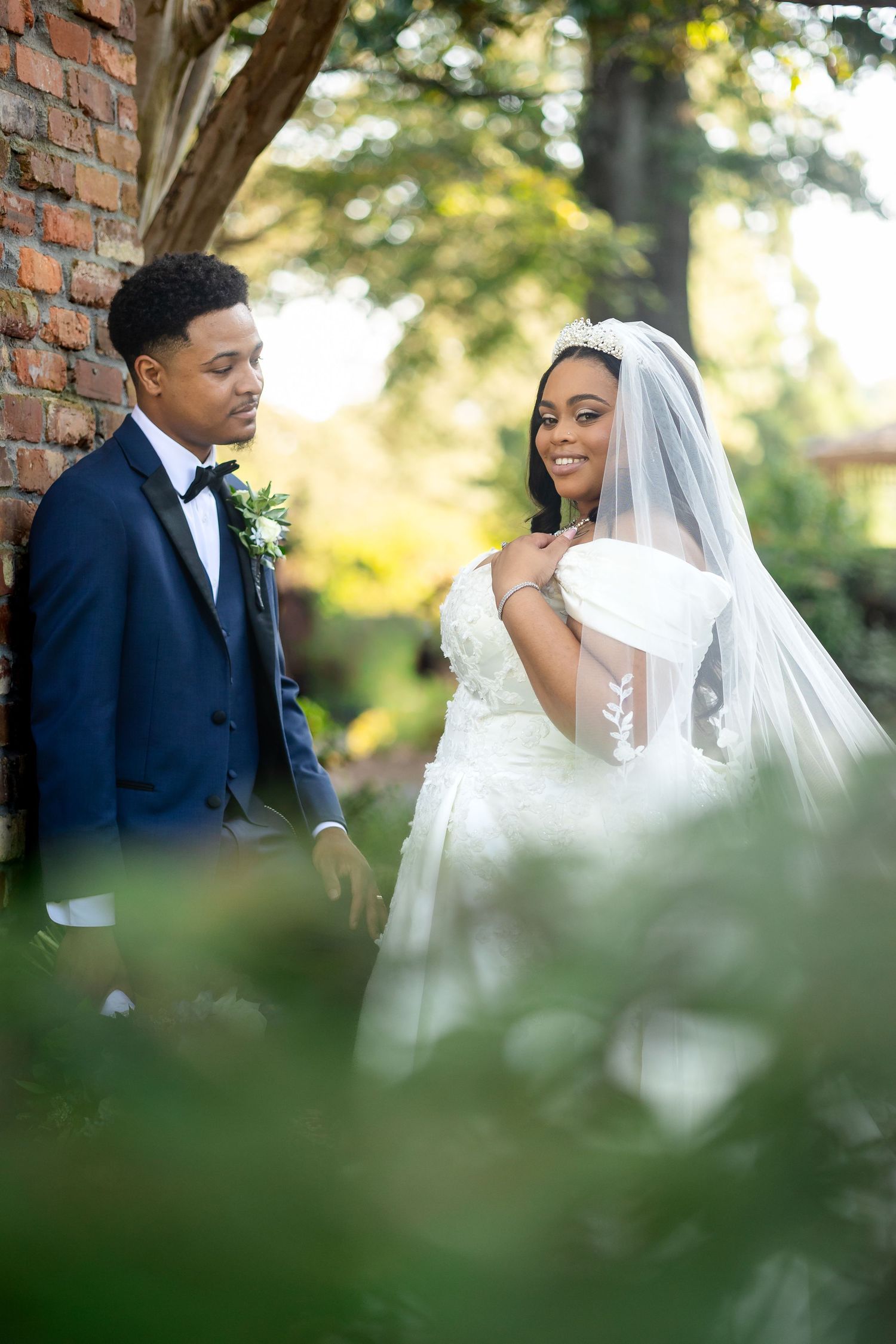 A bride in a white gown and veil stands with a groom in a navy suit near a brick wall surrounded by lush greenery.