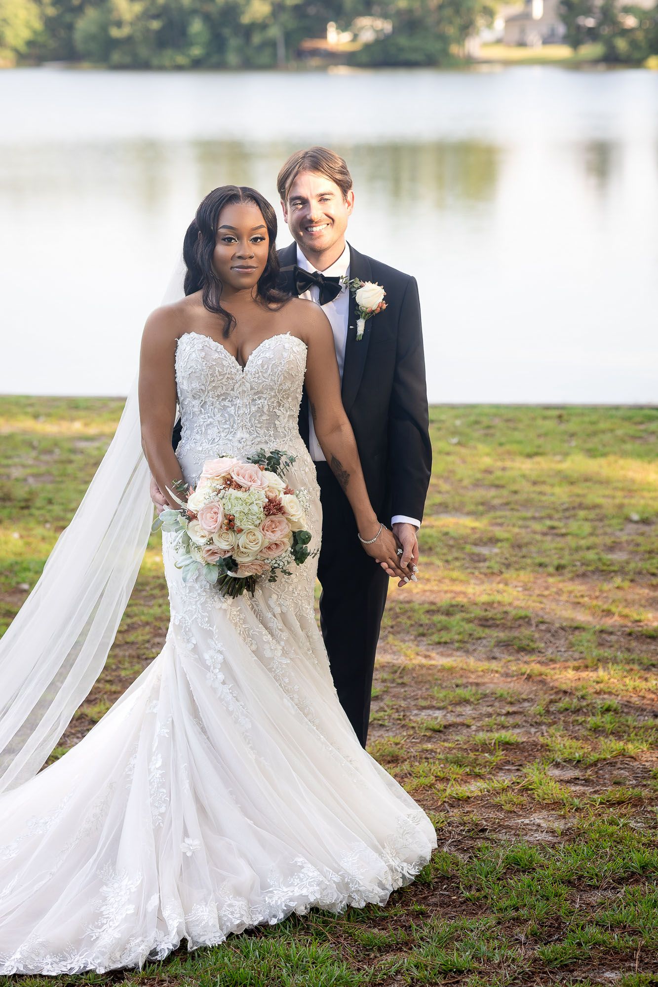 Interracial couple posing by a lake on their wedding day, bride in lace mermaid gown holding bouquet.