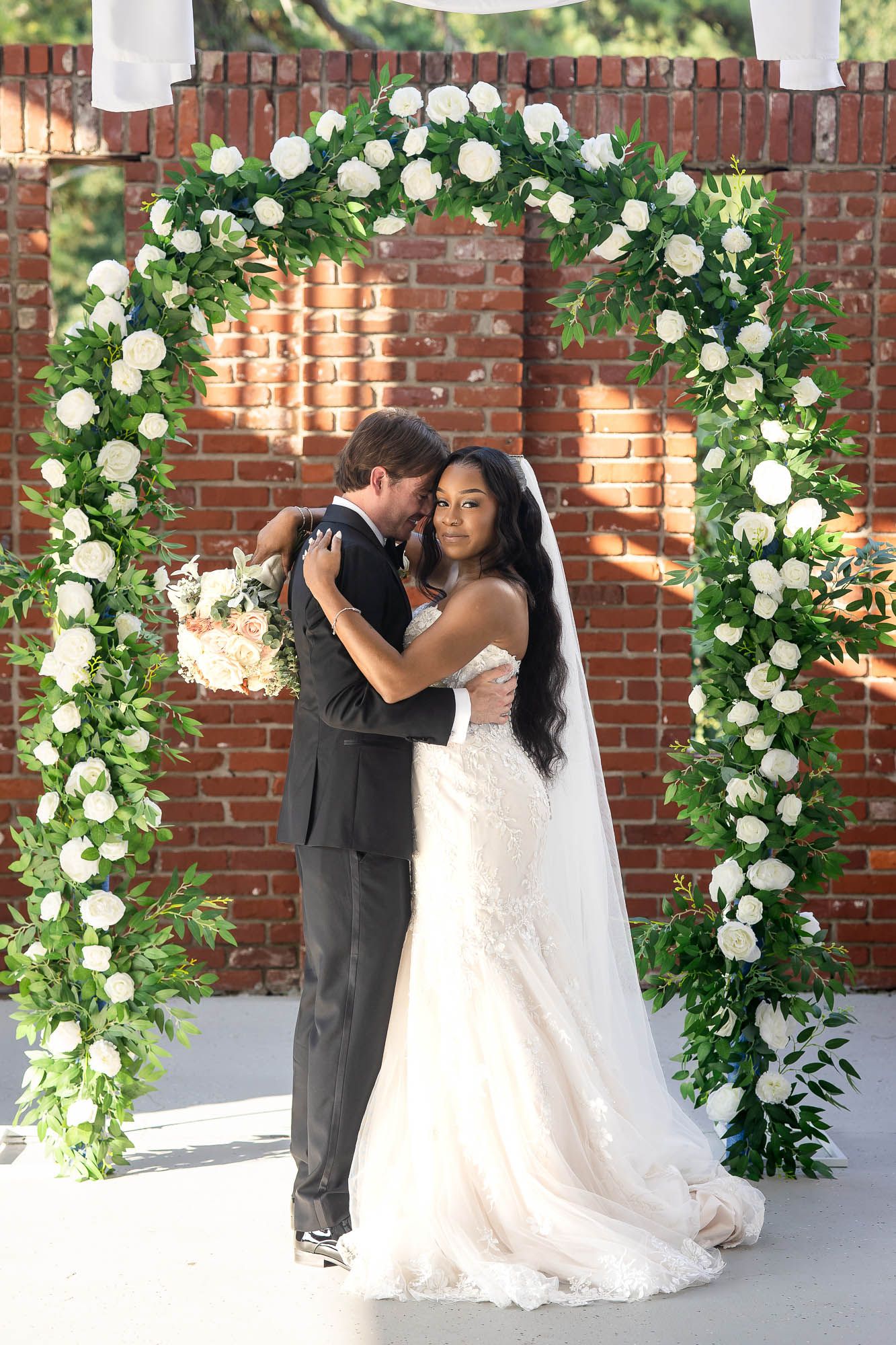 Bride and groom embrace under a floral arch adorned with white roses and greenery against a brick wall backdrop.