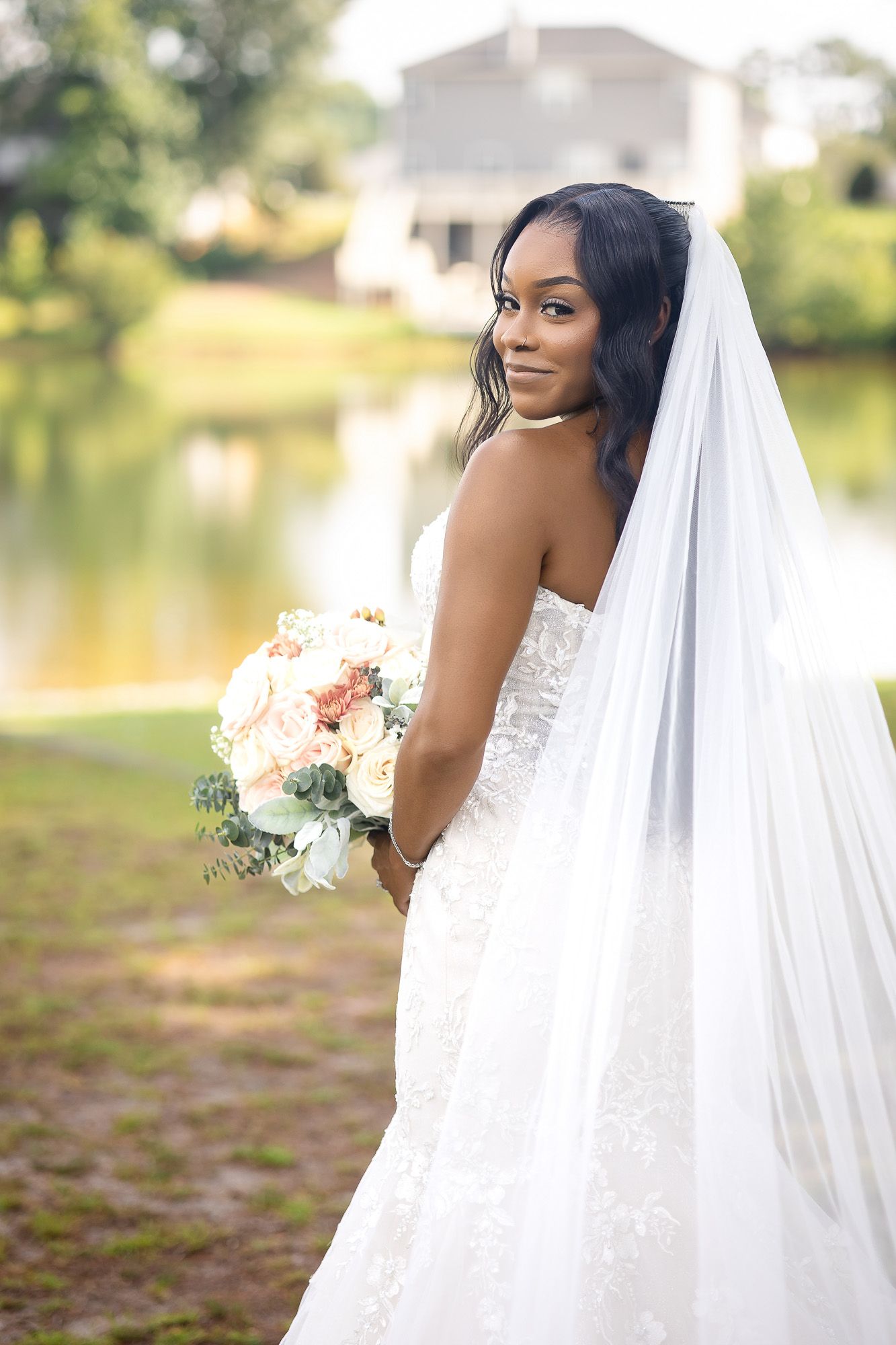 Elegant Black bride in lace gown and veil holding bouquet by lakeside.