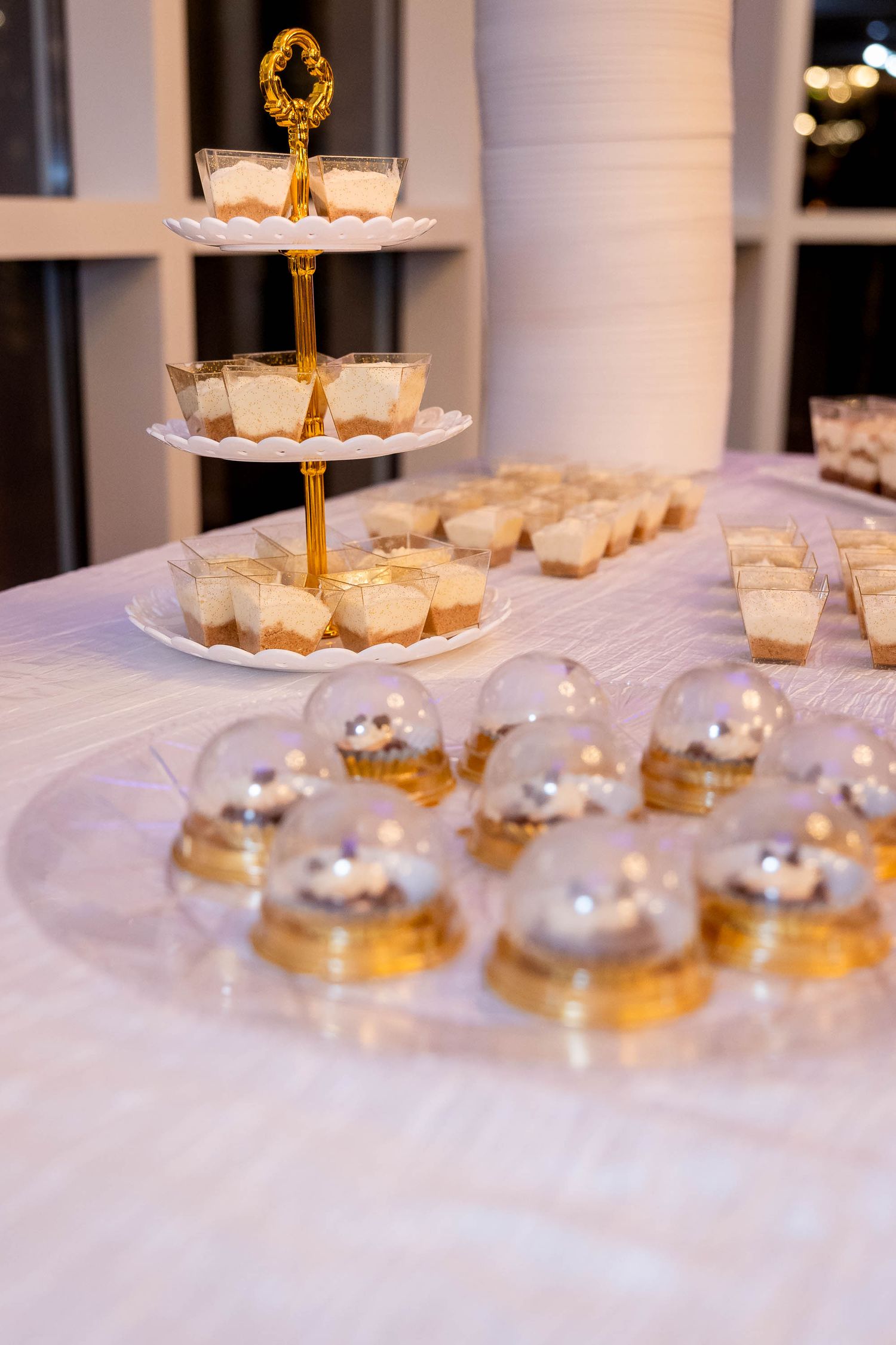 Elegant dessert table with gold tiered stand, mini cheesecakes, and dome-covered cupcakes at a formal event.