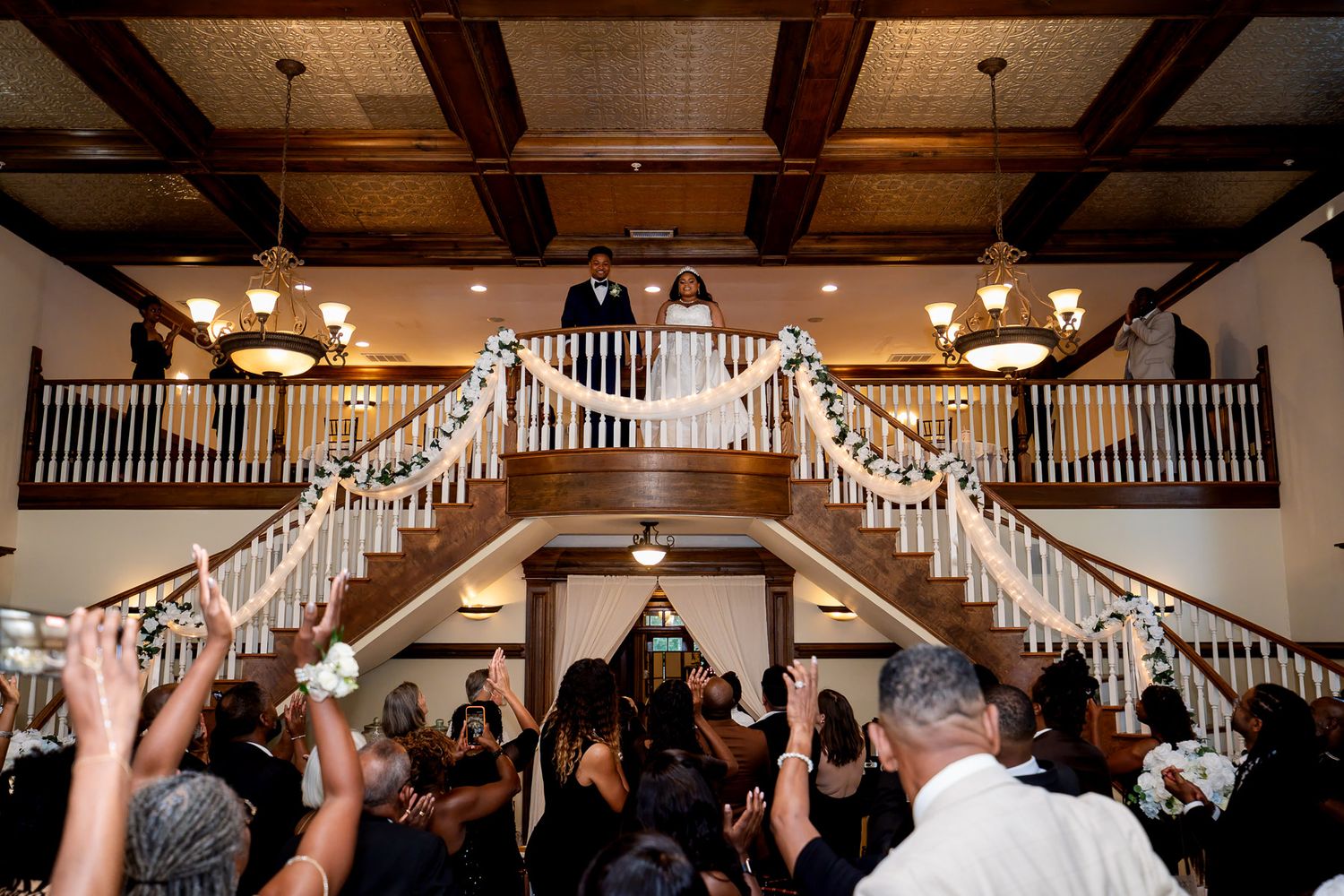 Bride and groom stand on elegant staircase balcony as wedding guests celebrate below in a grand reception hall.