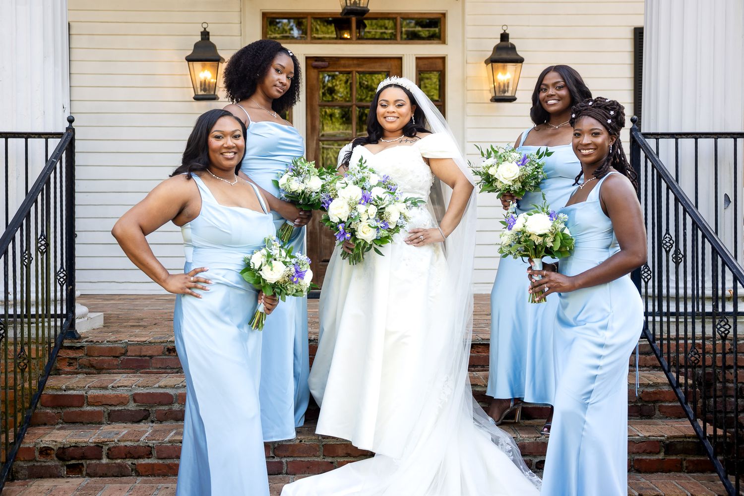 Bride in white gown poses with three bridesmaids in light blue dresses holding bouquets on elegant venue steps.