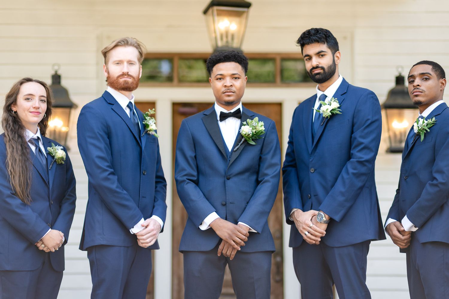 Groom in navy blue suit with bow tie stands with four groomsmen wearing matching suits and boutonnieres outdoors.