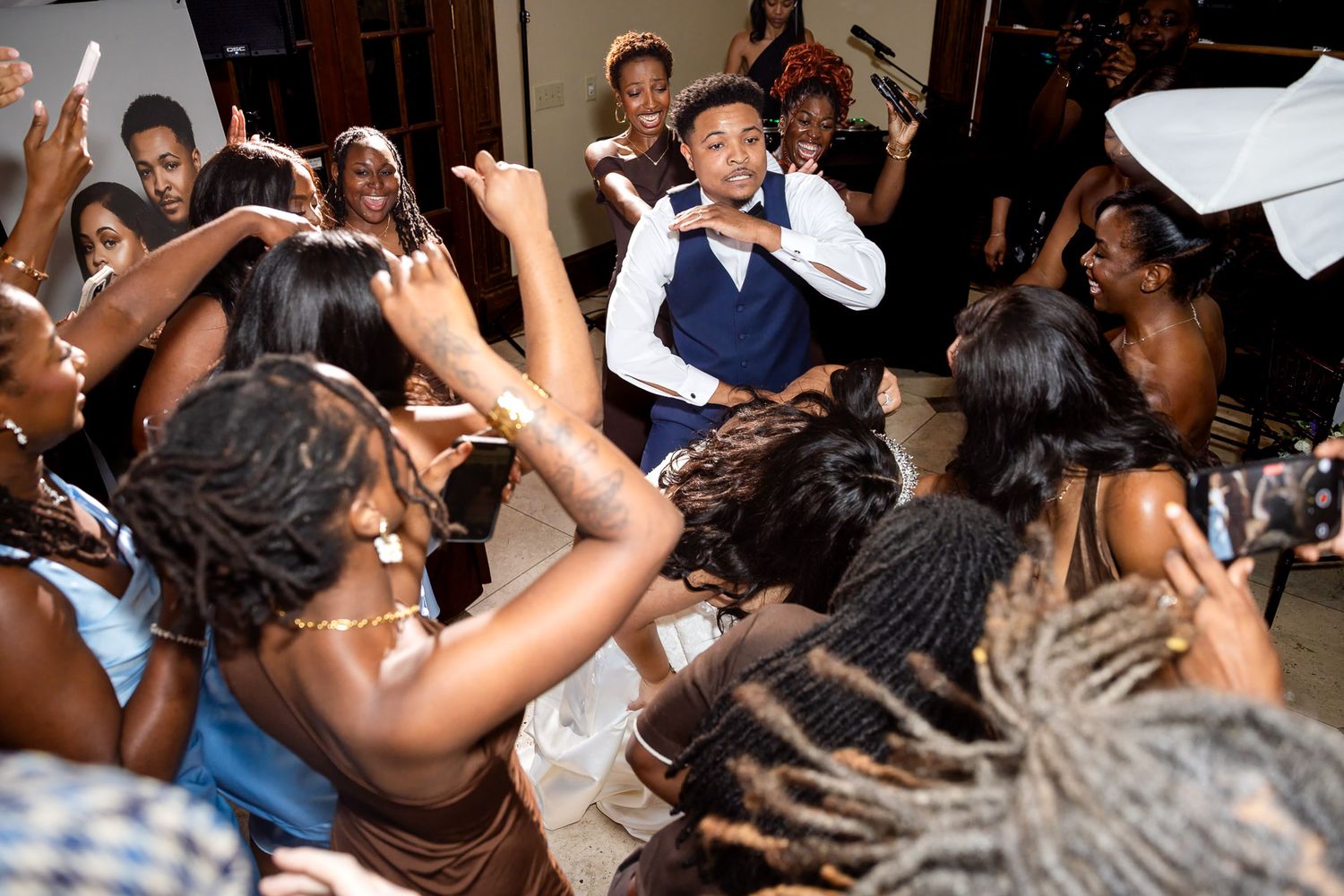 A lively wedding reception dance floor with guests surrounding a joyful groom in a blue vest celebrating together.
