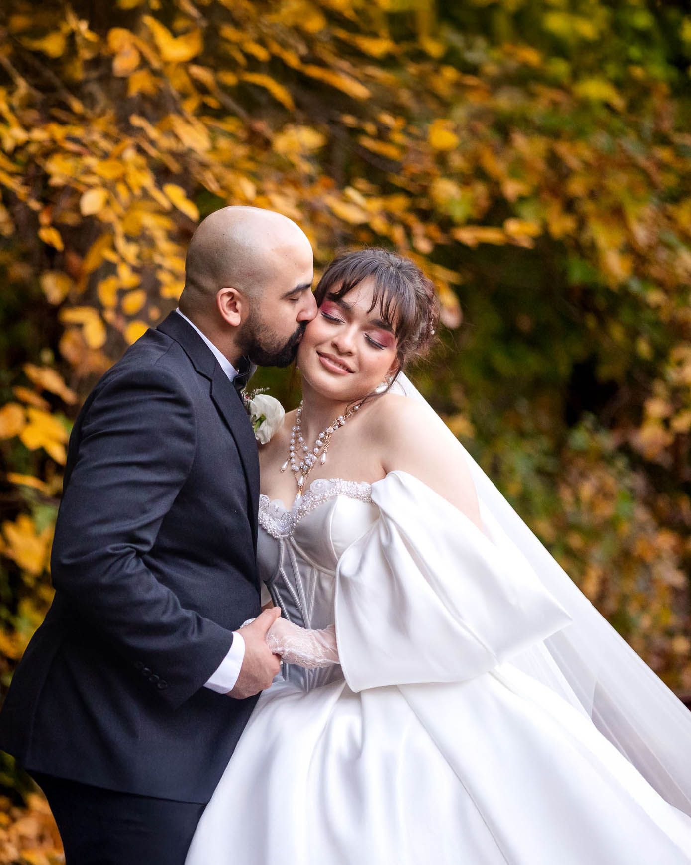 Bride and groom share a tender kiss among golden autumn leaves, she in a white ballgown, he in a dark suit.