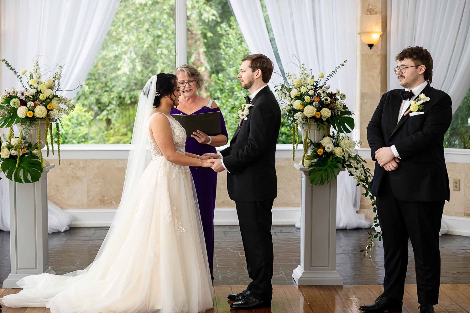Bride and groom exchange vows at elegant indoor wedding ceremony with floral arrangements and officiant.