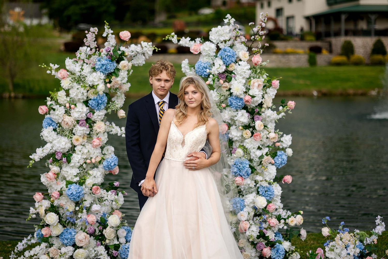 Wedding couple poses under elegant floral arch with white and blue flowers beside tranquil lake.