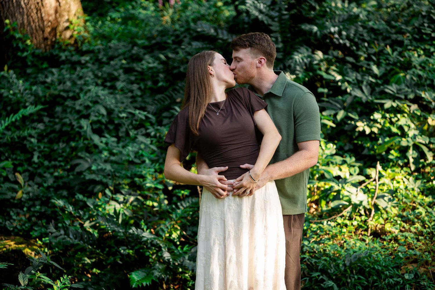 Couple sharing a tender kiss outdoors, hands on baby bump, surrounded by lush green foliage in a maternity photo.