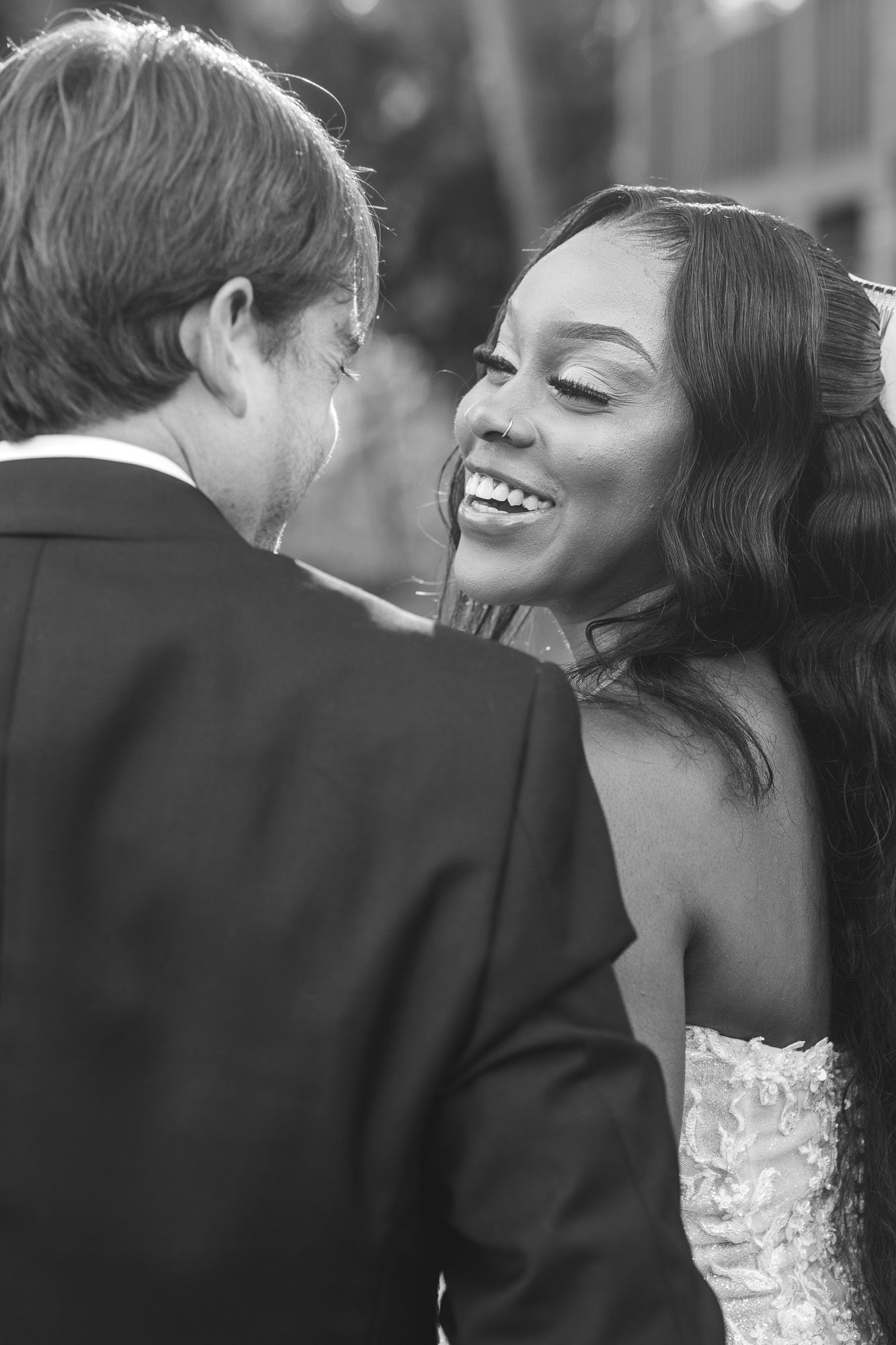 Black and white photo of a smiling bride in lace gown leaning toward groom in tuxedo at wedding ceremony.