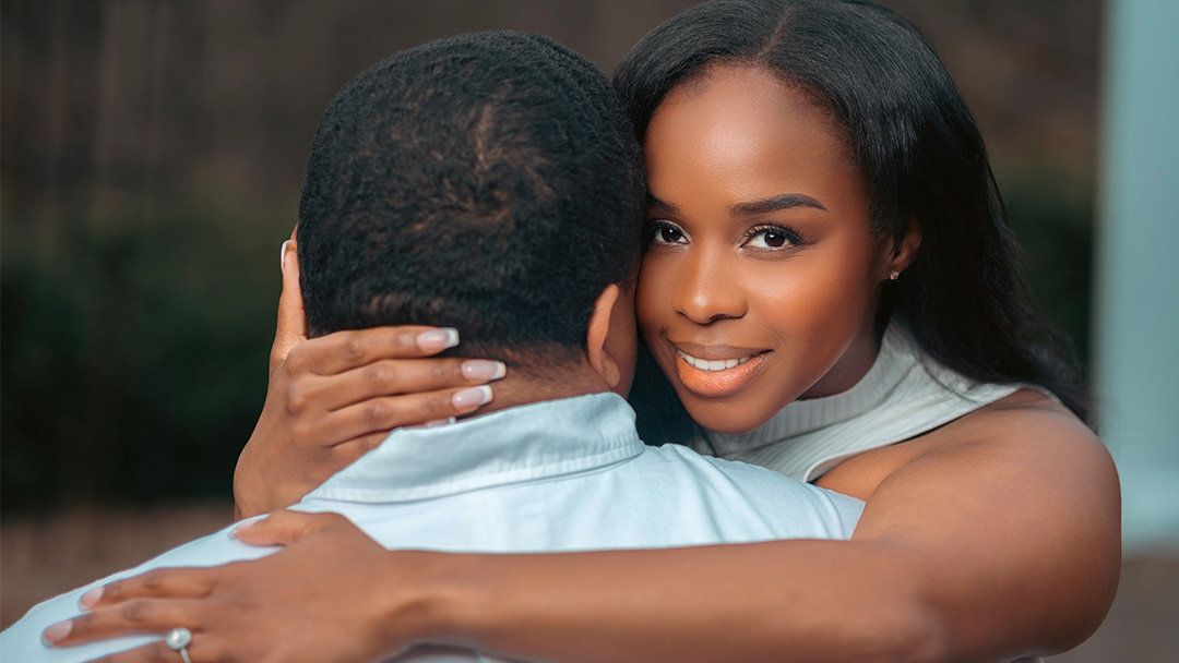 A young woman smiles warmly while embracing a man outdoors, wearing a light blue outfit and a ring.