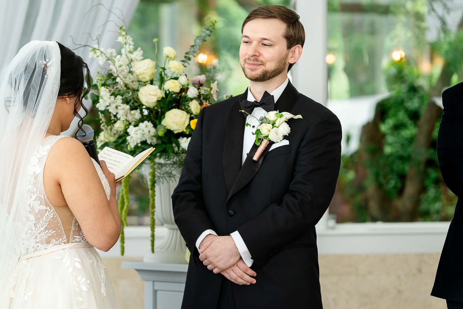 Groom in black tuxedo smiling at bride during wedding ceremony with white floral arrangements in background.