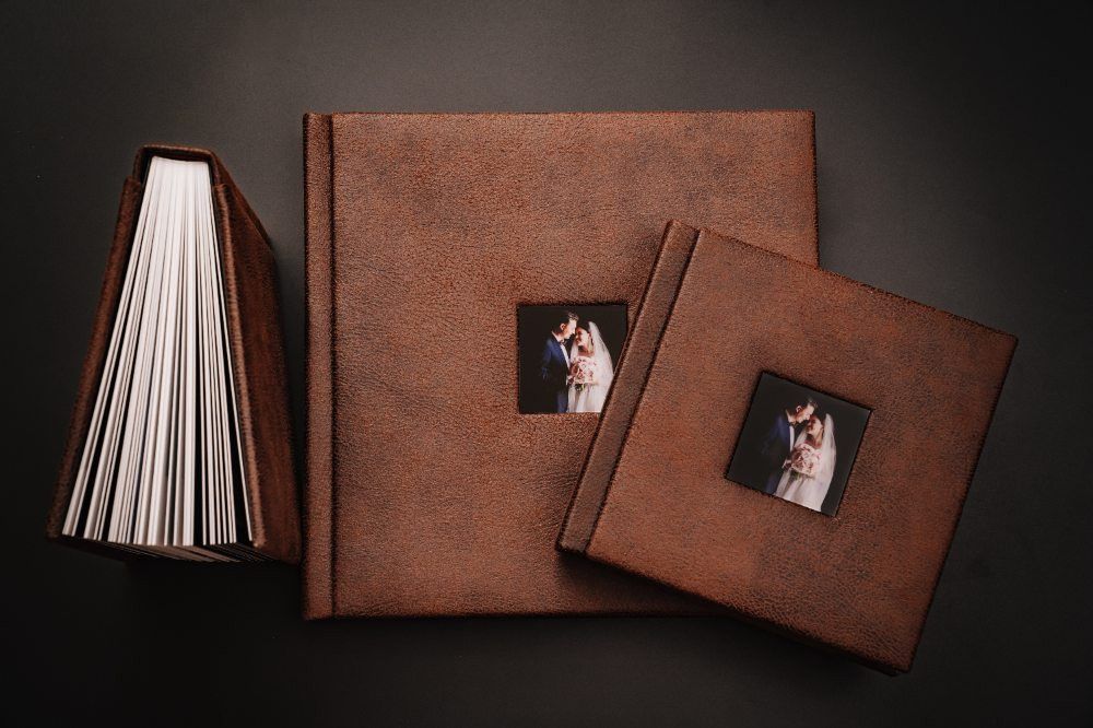 Three brown leather photo albums with wedding couple image windows displayed on dark background.