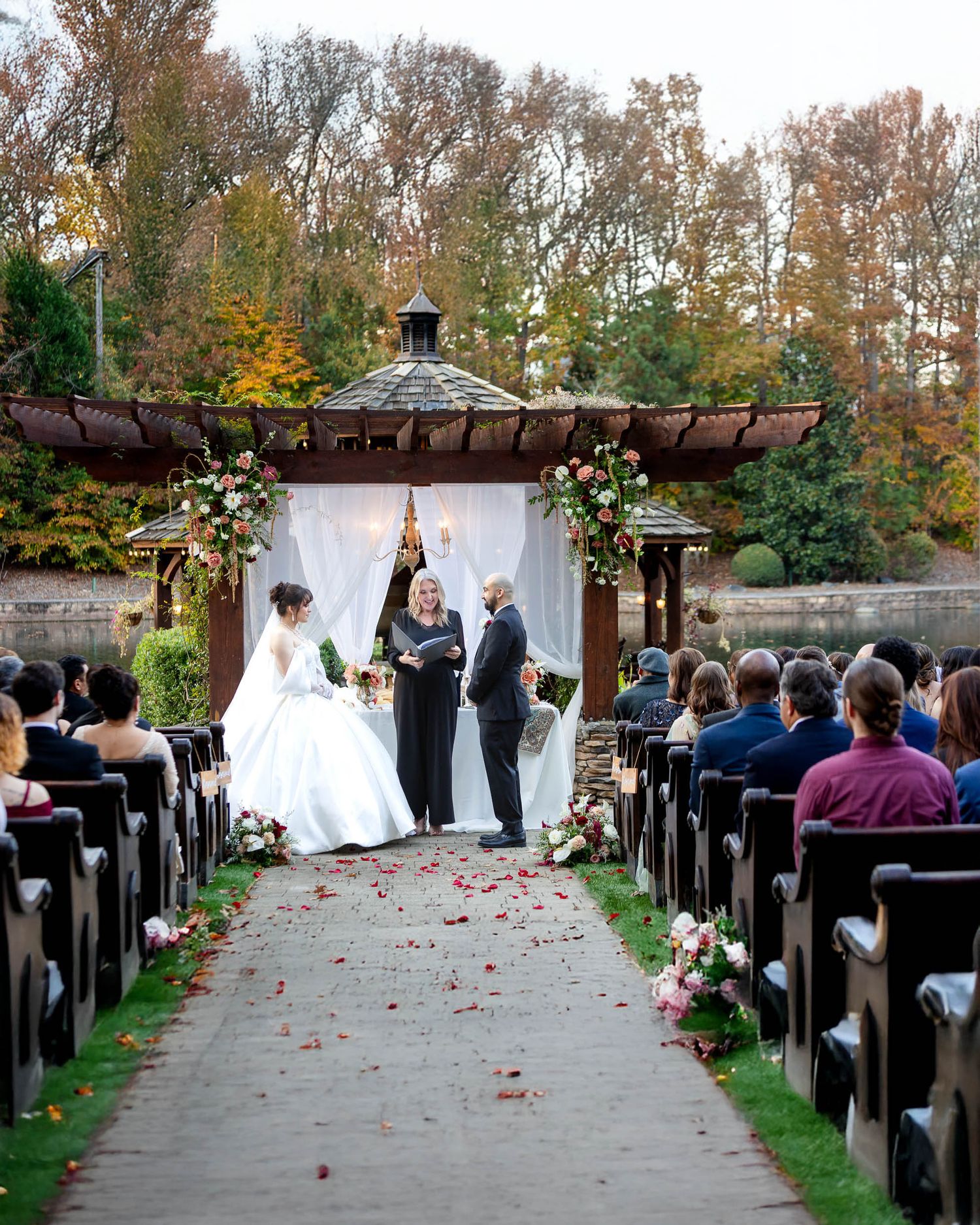 Outdoor wedding ceremony at a gazebo with floral arch, rose petal aisle, and guests seated in wooden pews amid autumn foliage.