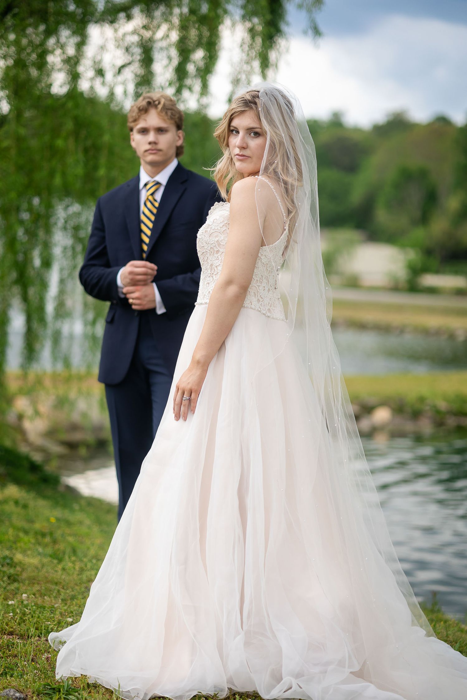 A couple poses for wedding photos by a serene lake with willow trees, bride wearing a flowing white dress and veil.