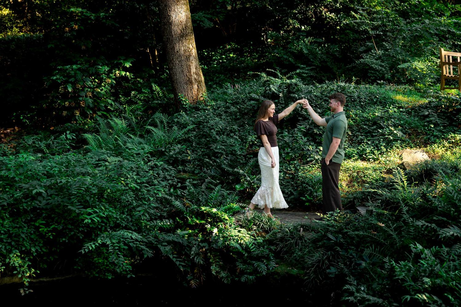 Couple dancing together on a wooden bridge surrounded by lush green ferns in a forest setting.