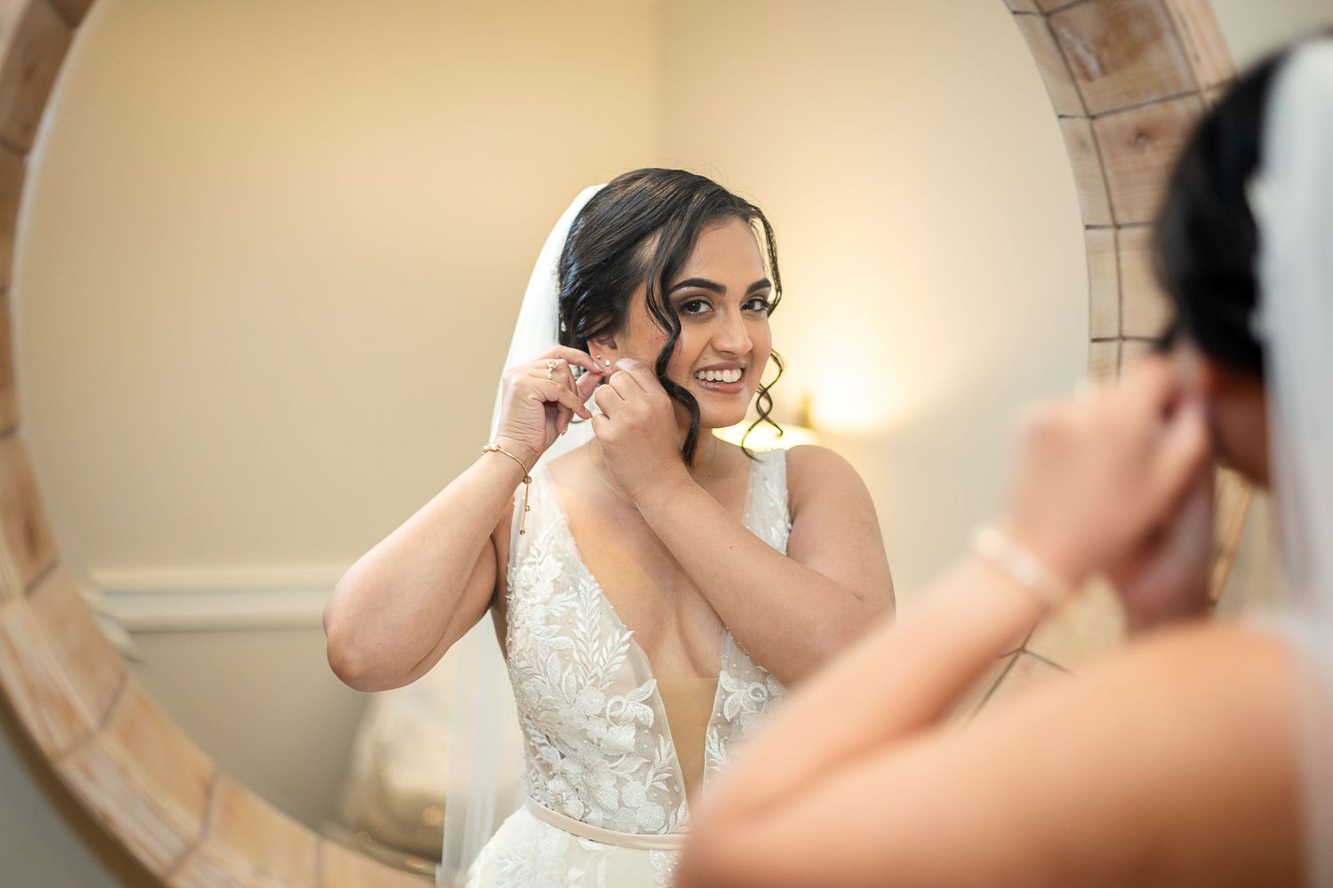Bride in lace wedding dress adjusting earring while smiling at her reflection in a round wooden mirror.