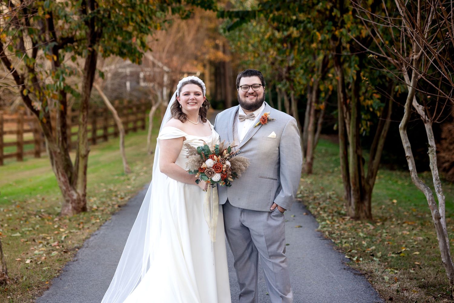 Bride in white gown holding bouquet stands beside groom in gray suit on tree-lined path at outdoor fall wedding.