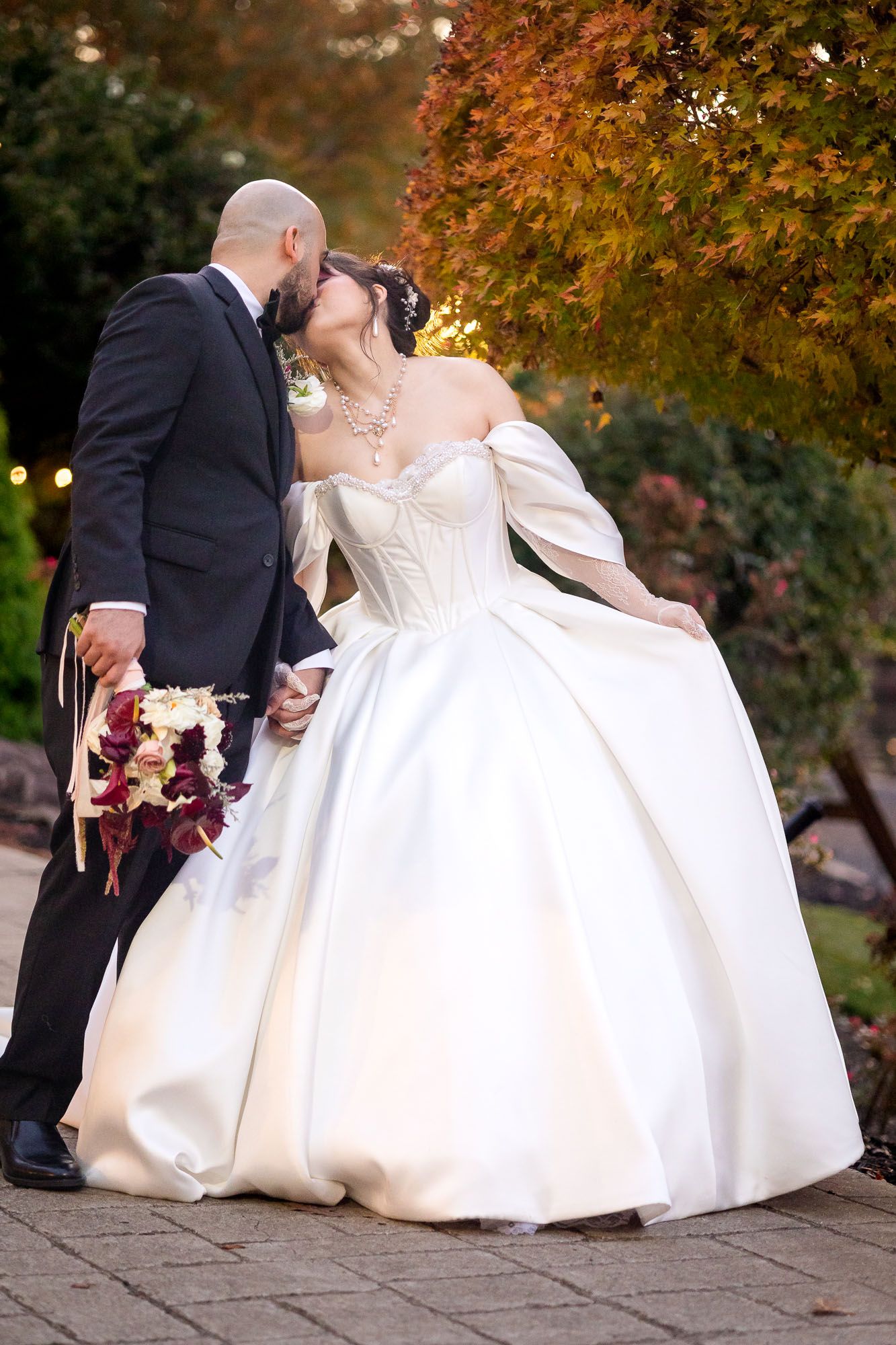 Bride and groom sharing a romantic kiss outdoors, wearing elegant wedding attire with colorful autumn foliage in the background.