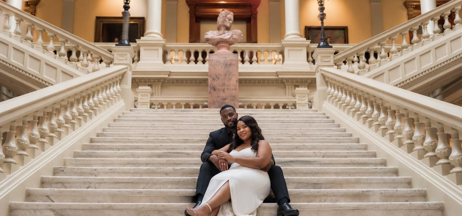 Couple in formal attire sitting on grand marble staircase with classical statue backdrop for elegant engagement photos.