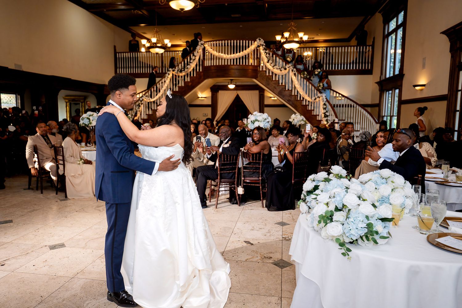 Bride and groom share first dance at elegant wedding reception with floral arrangements and guests seated nearby.