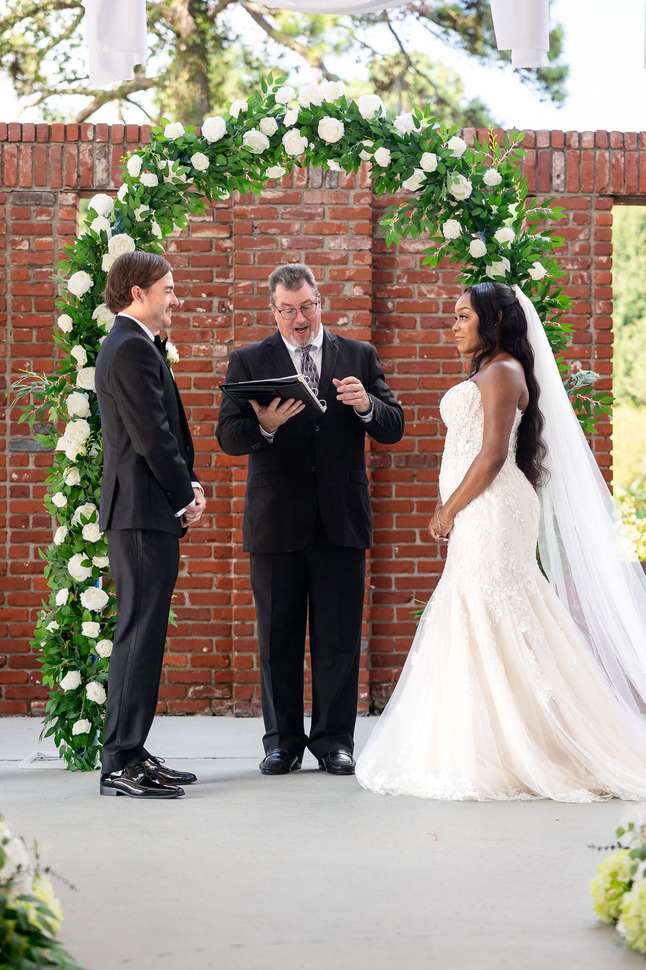 Bride and groom exchange vows under a floral arch with white roses and greenery against a brick wall.
