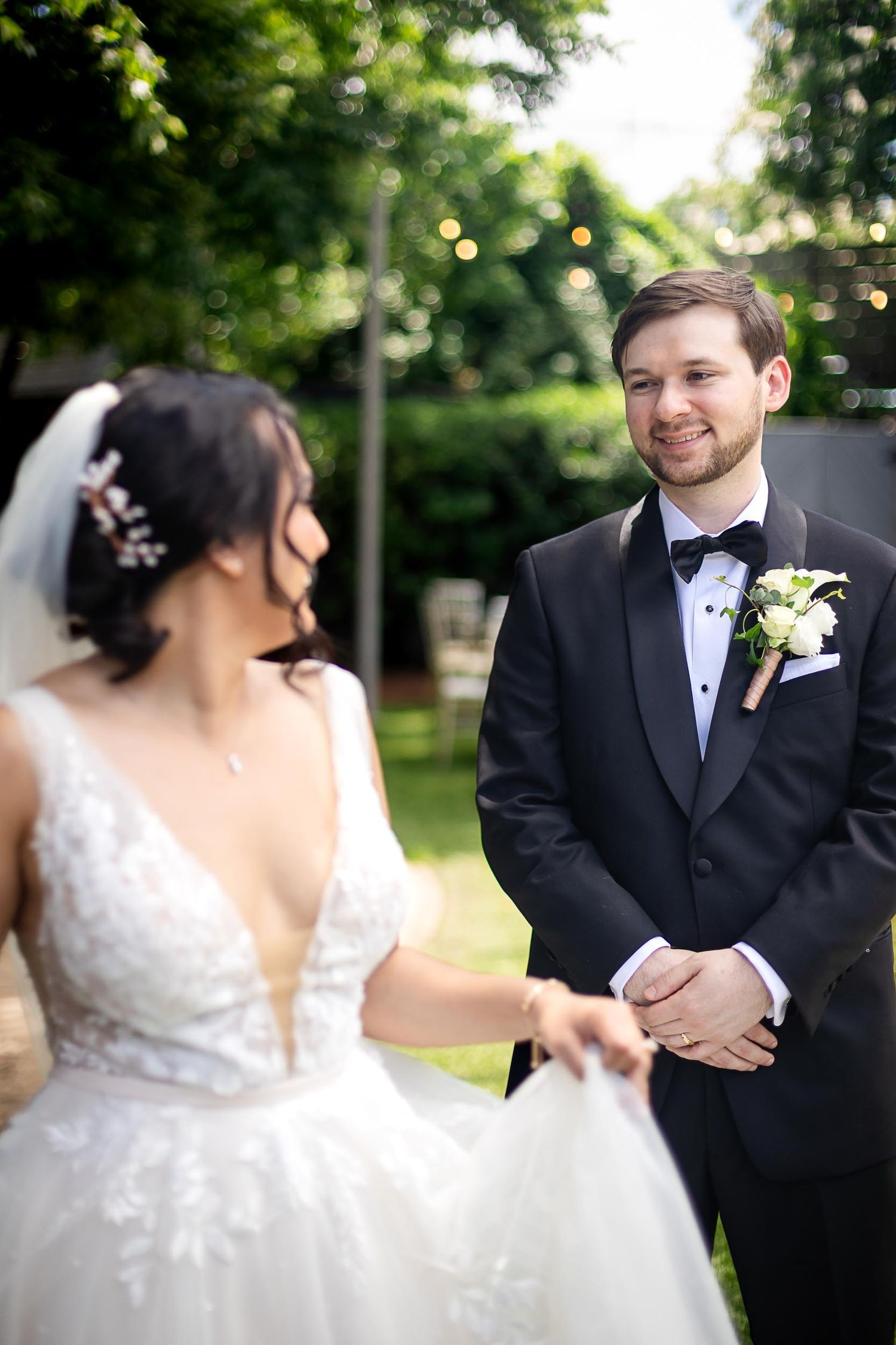 Groom in black tuxedo smiling at bride in white lace gown during outdoor wedding ceremony.