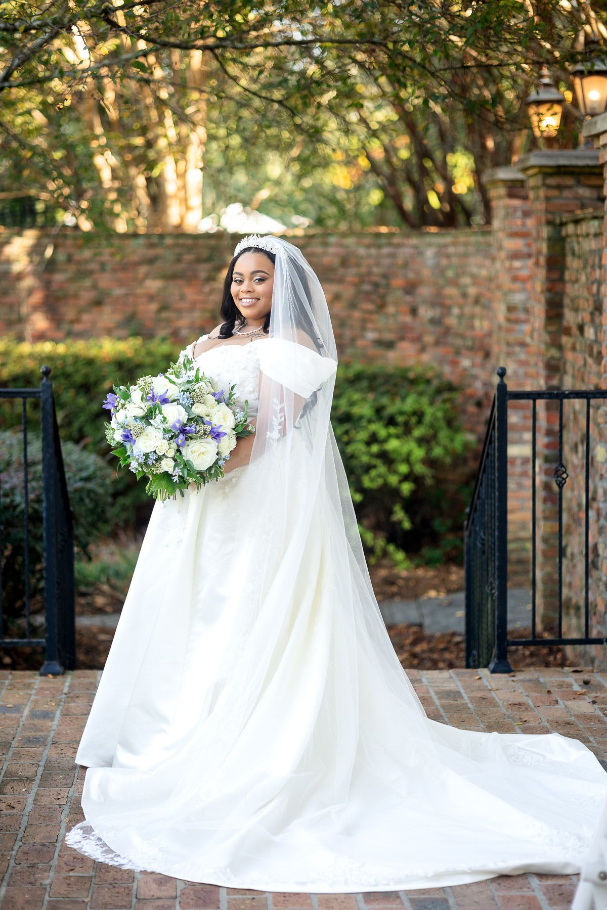 Smiling Black bride in white off-shoulder gown with veil and floral crown holds blue and white bouquet on brick patio.