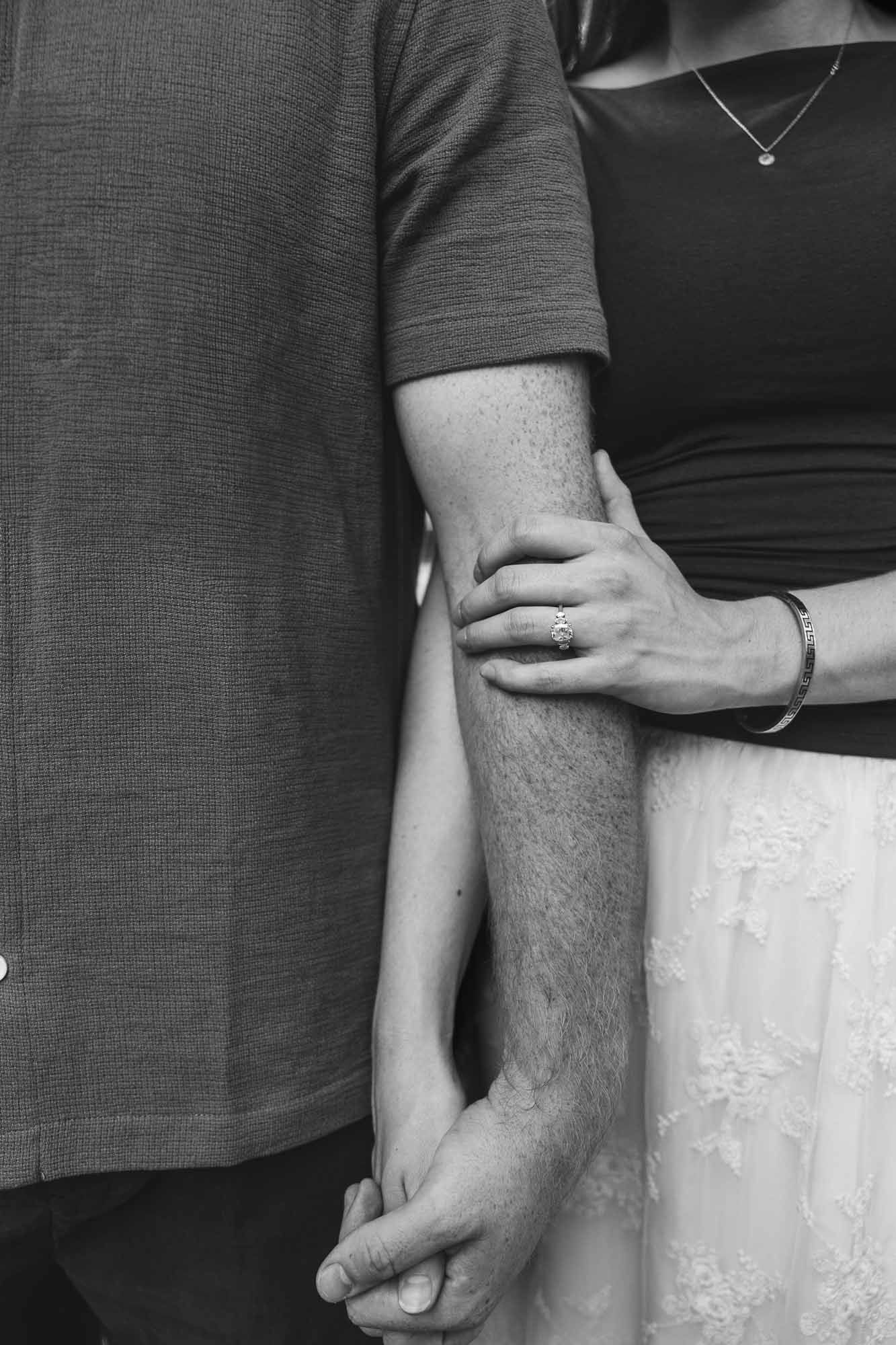 Black and white photo of a couple holding hands, woman wearing engagement ring and bracelet, man in grey shirt.