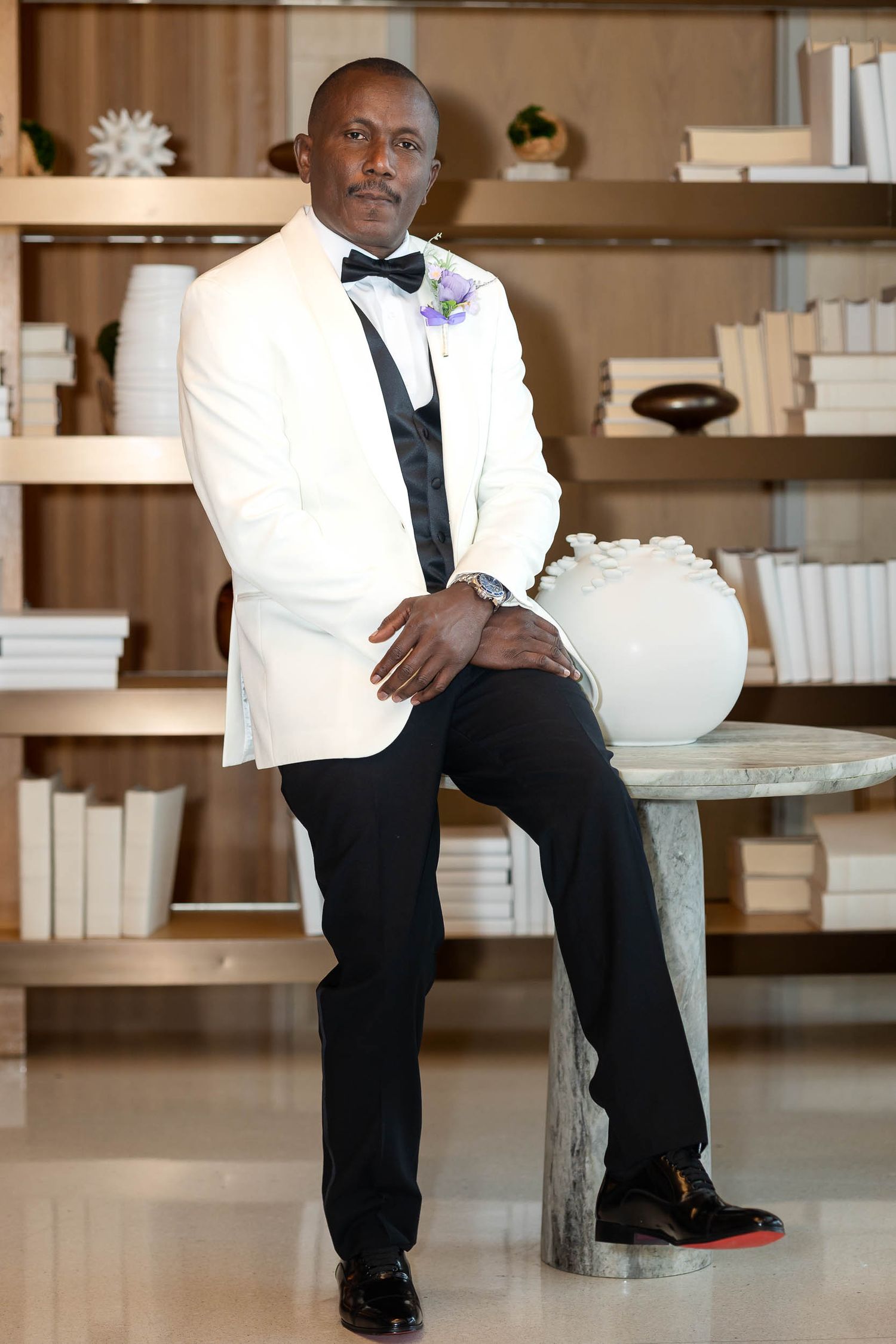 Groom in white tuxedo jacket and black bow tie sits confidently in front of a styled bookshelf.