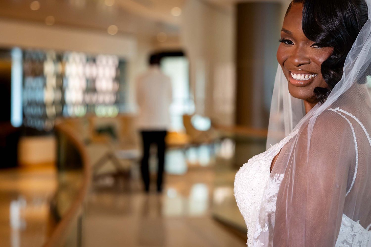 Smiling Black bride in white lace wedding dress and veil, with groom standing in background at elegant venue.