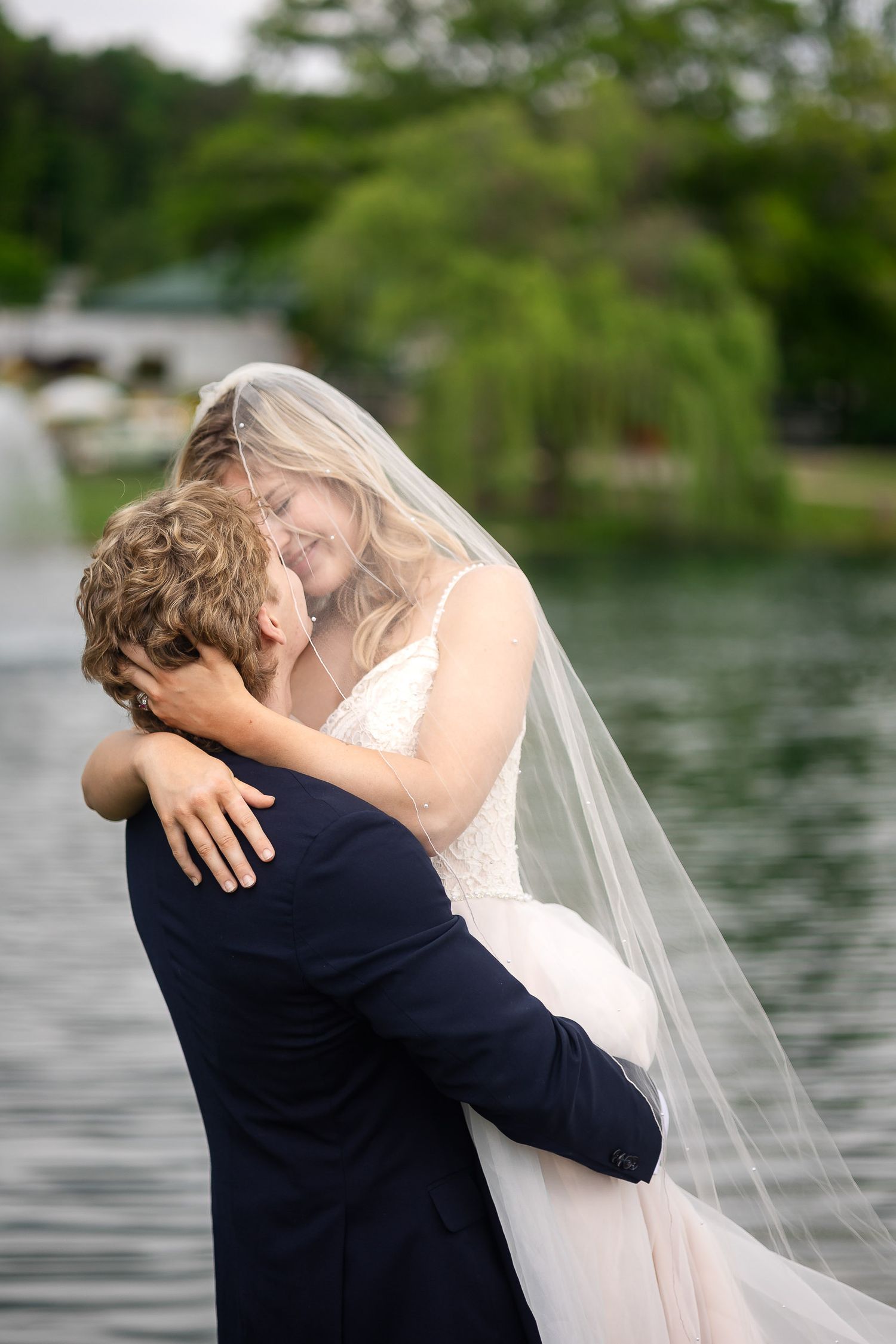 A romantic wedding couple embraces by a serene lakeside with trees in the background.