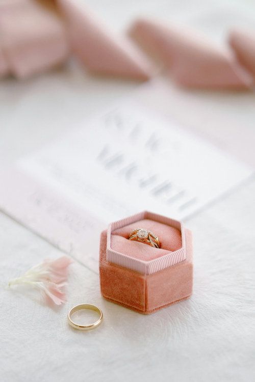 Gold diamond engagement ring in a pink velvet hexagon box with a wedding band and invitation on white fur.