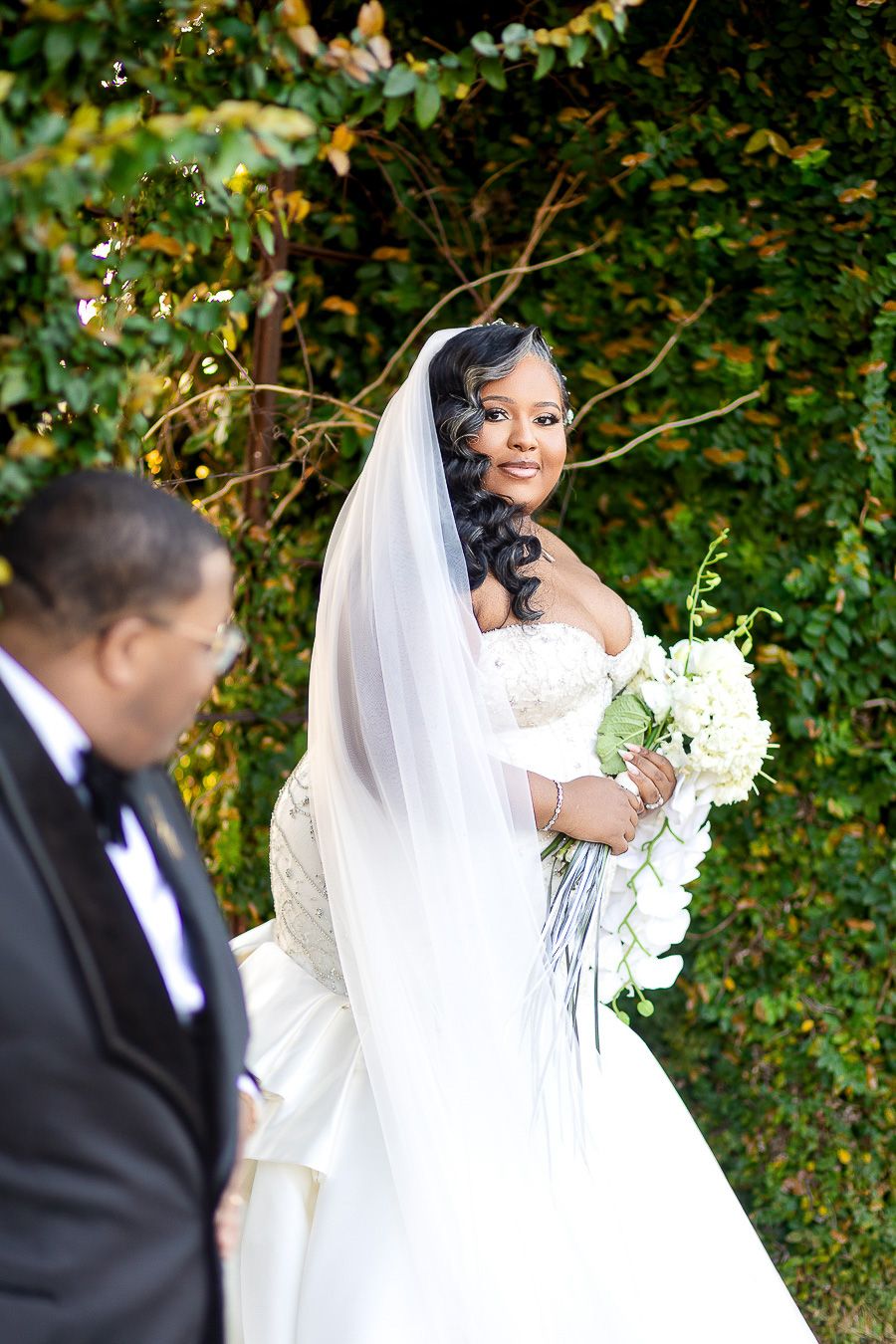 A radiant Black bride in a white gown and veil holds a bouquet, smiling outdoors near lush greenery at a wedding at Bibb Mill Event Center