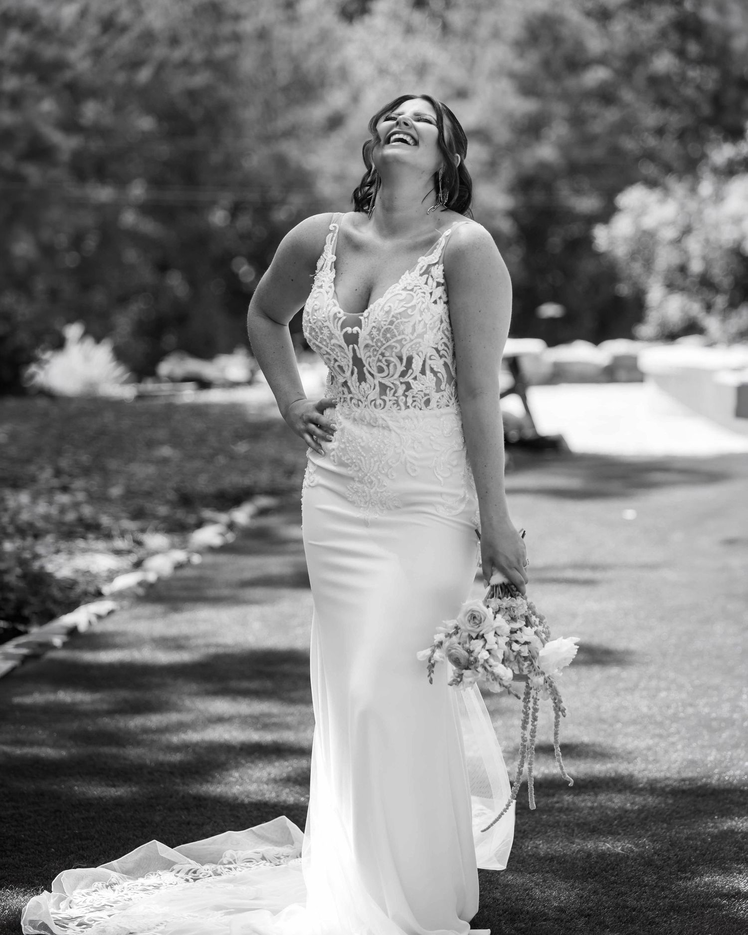 Bride in lace wedding gown laughing joyfully outdoors, holding bouquet, black and white photo.