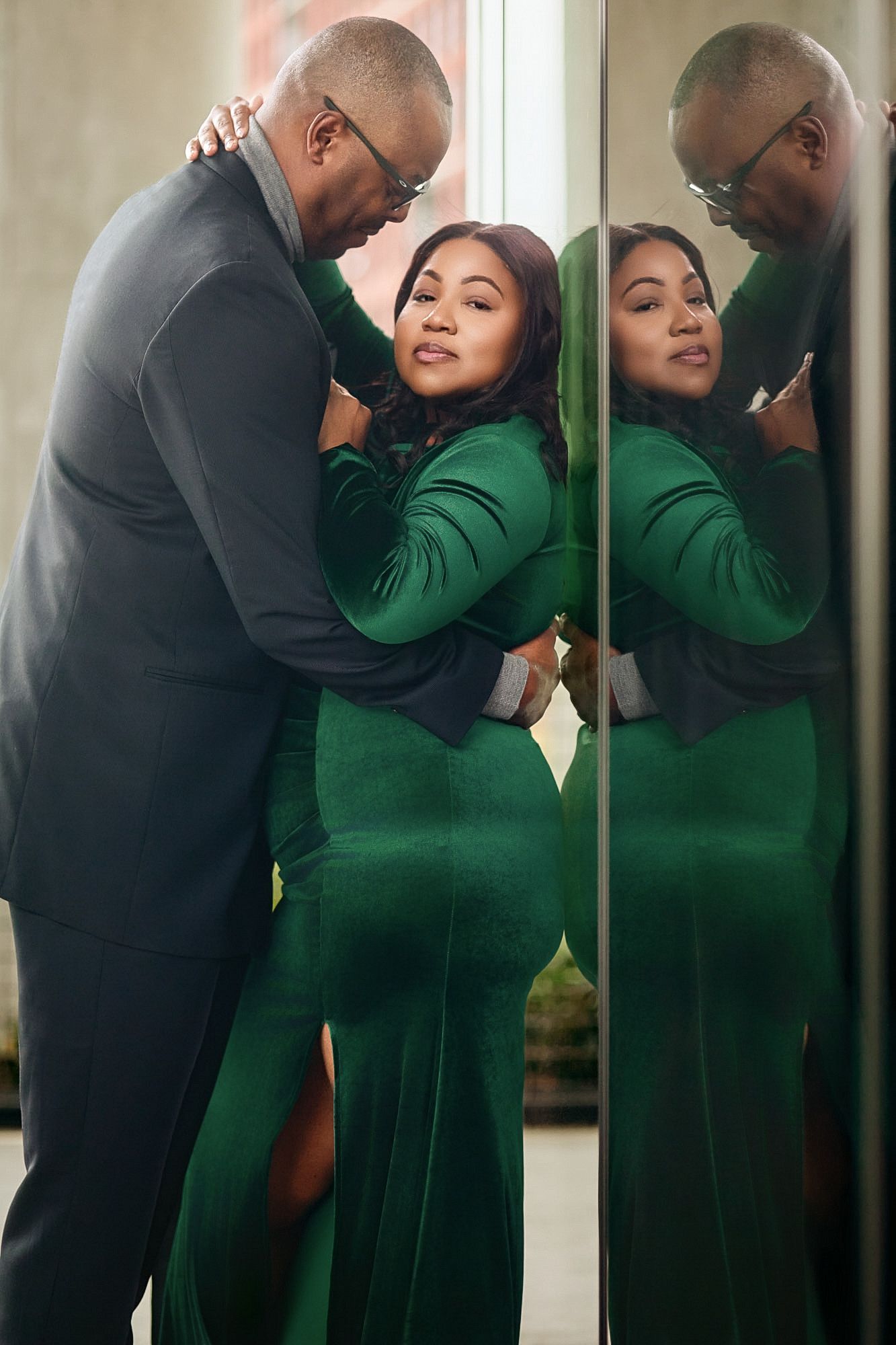 Couple embracing in green dress and gray suit, reflected in mirror, romantic portrait photography.