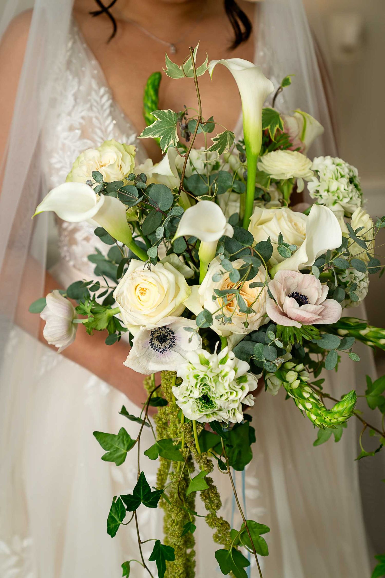 Bride holding a lush cascading wedding bouquet with white calla lilies, roses, anemones, and trailing greenery.