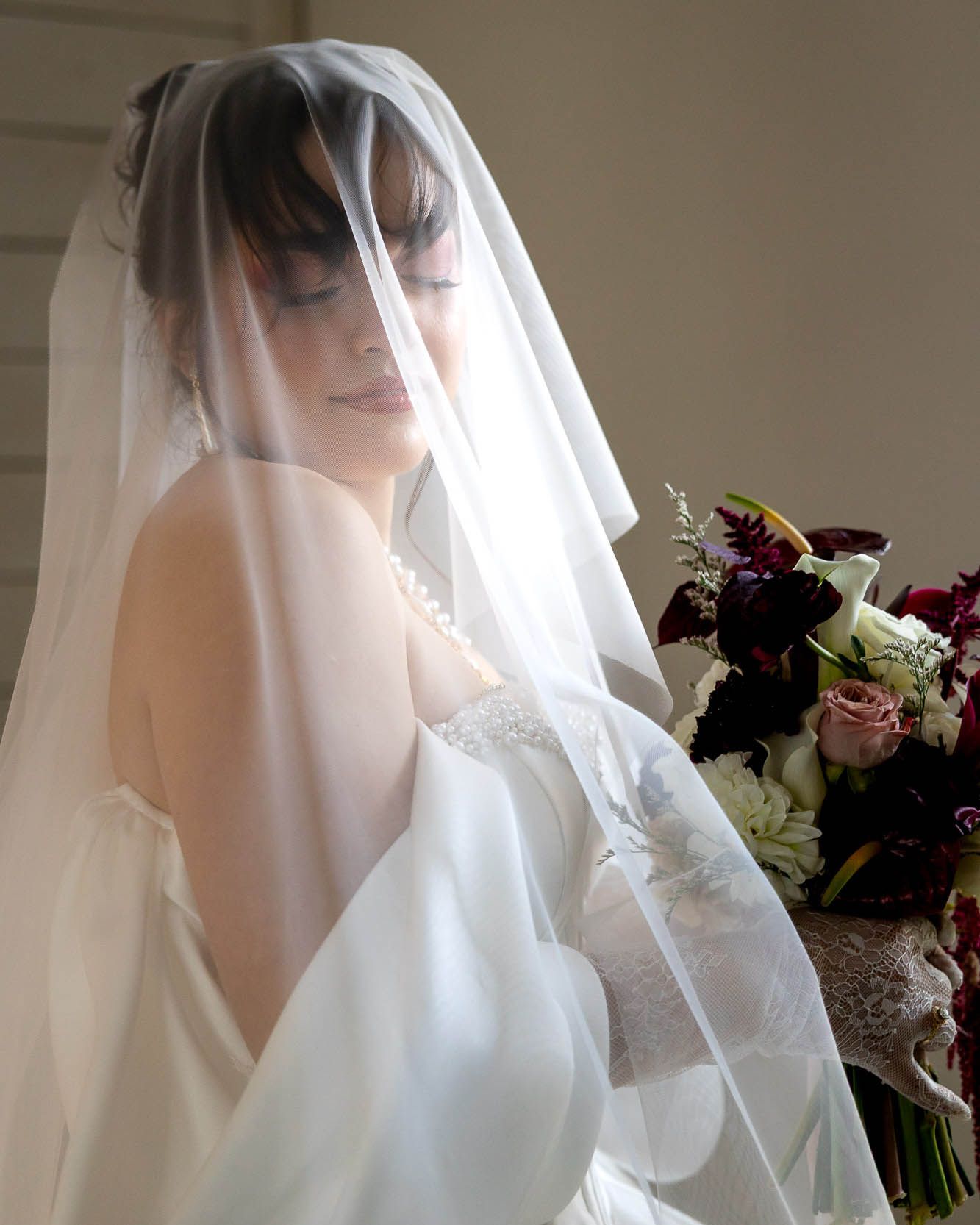 Bride in white wedding gown and sheer veil holds bouquet of burgundy and white flowers, eyes closed, serene expression.