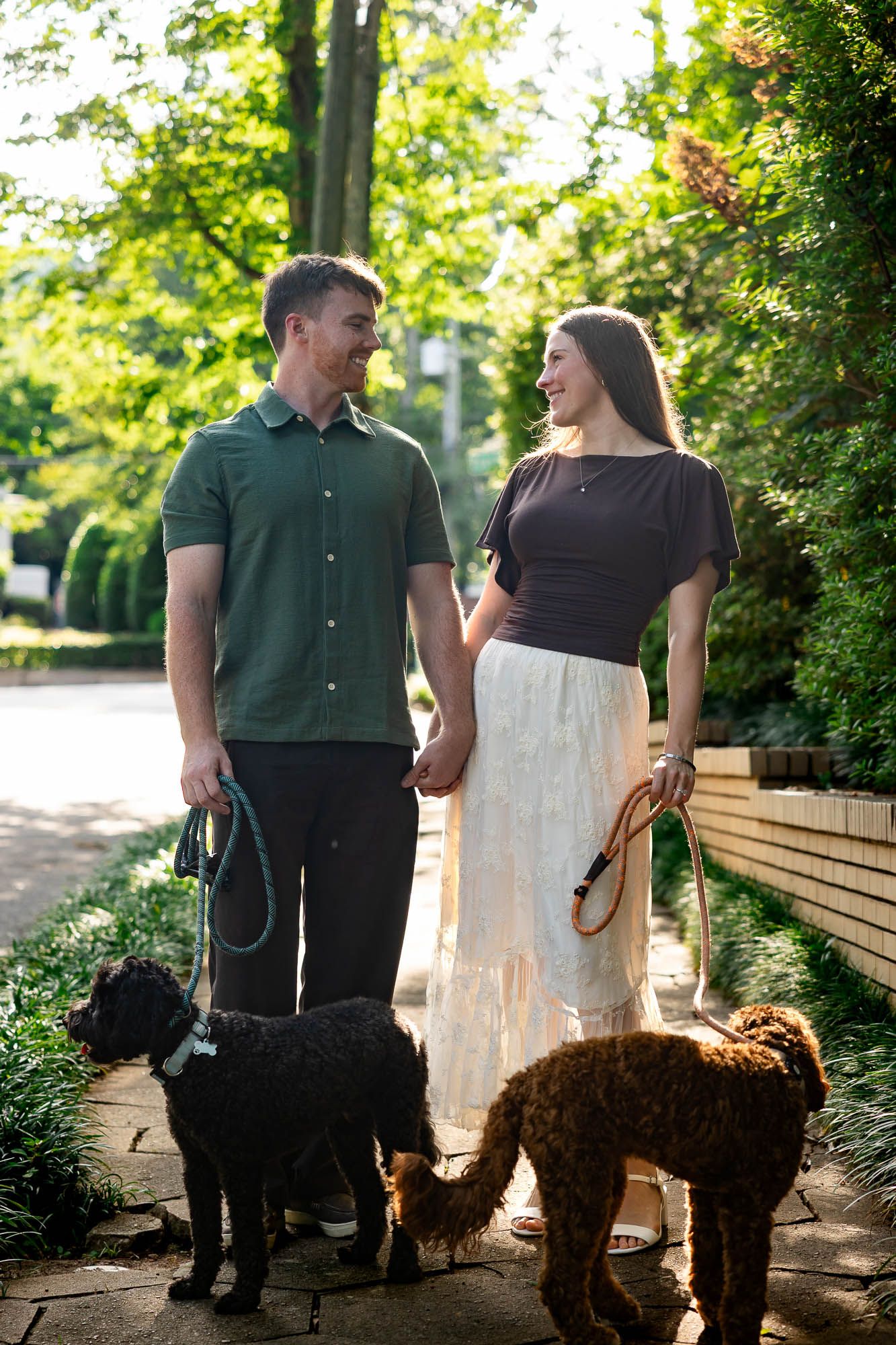 A smiling couple walks two doodle dogs on leashes along a sunlit garden path surrounded by lush green trees.