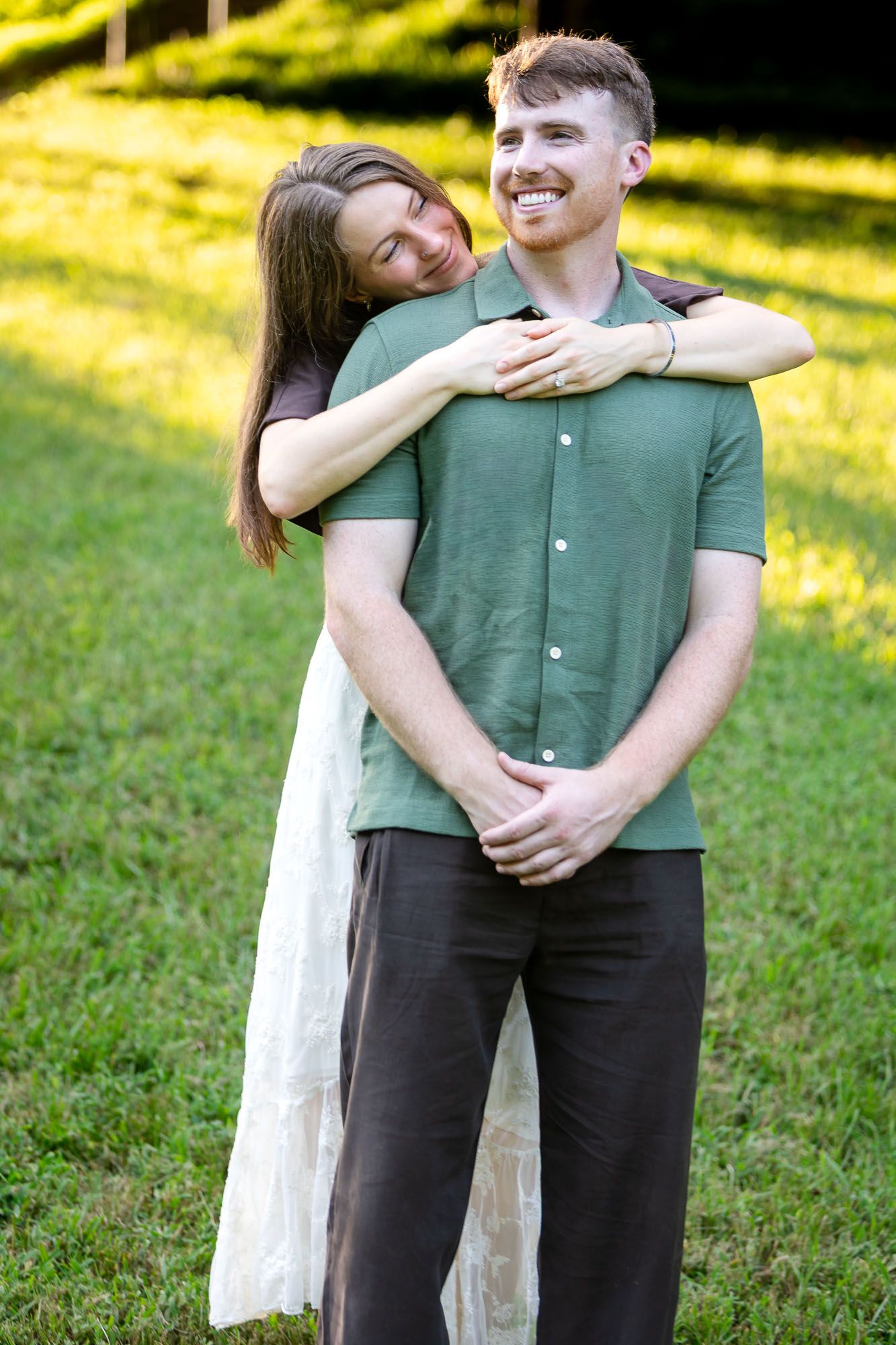 A smiling couple embraces outdoors on green grass, woman in white dress hugging man in green polo shirt.