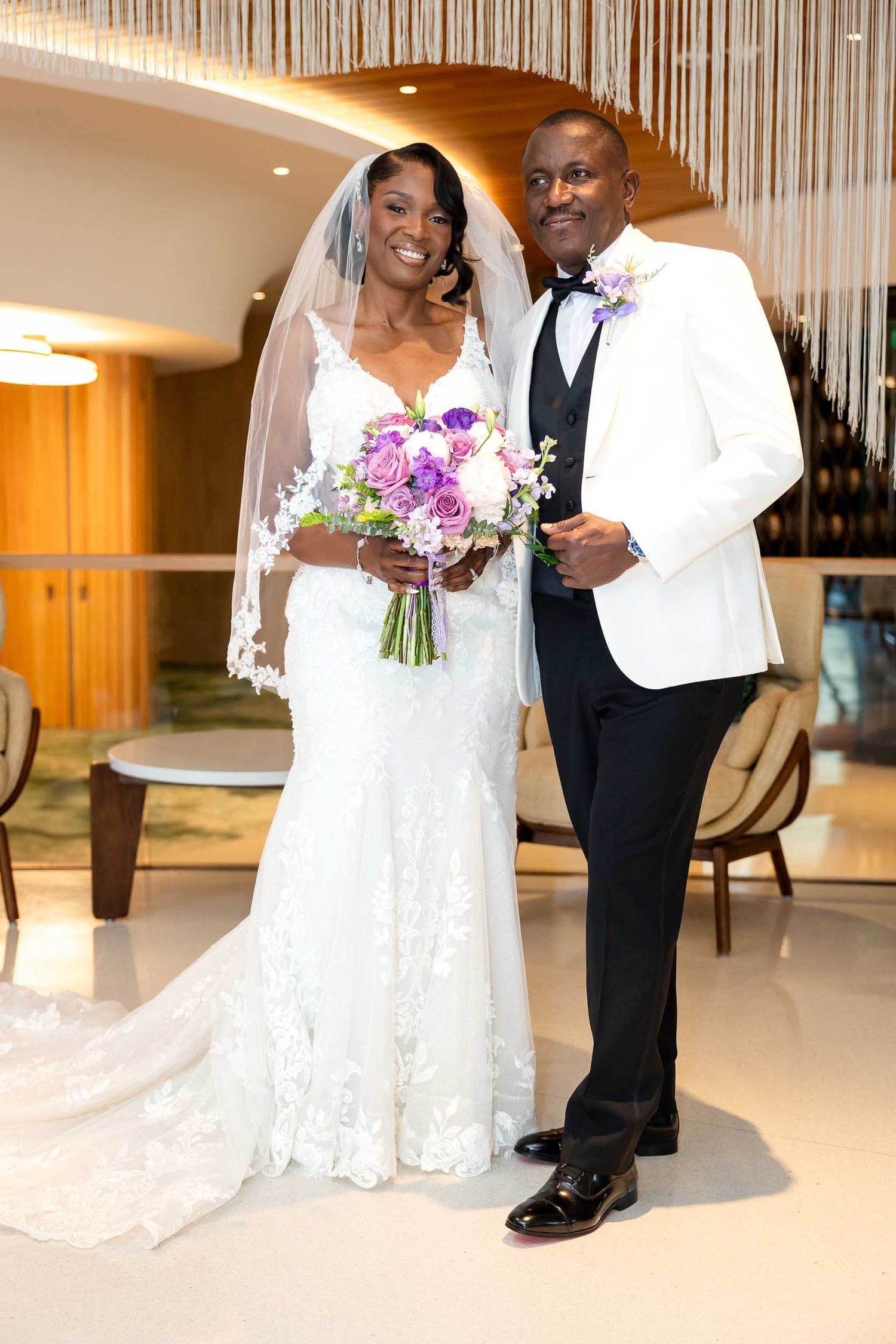 Bride in lace gown with veil holds purple bouquet beside groom in white tuxedo jacket at elegant wedding venue.