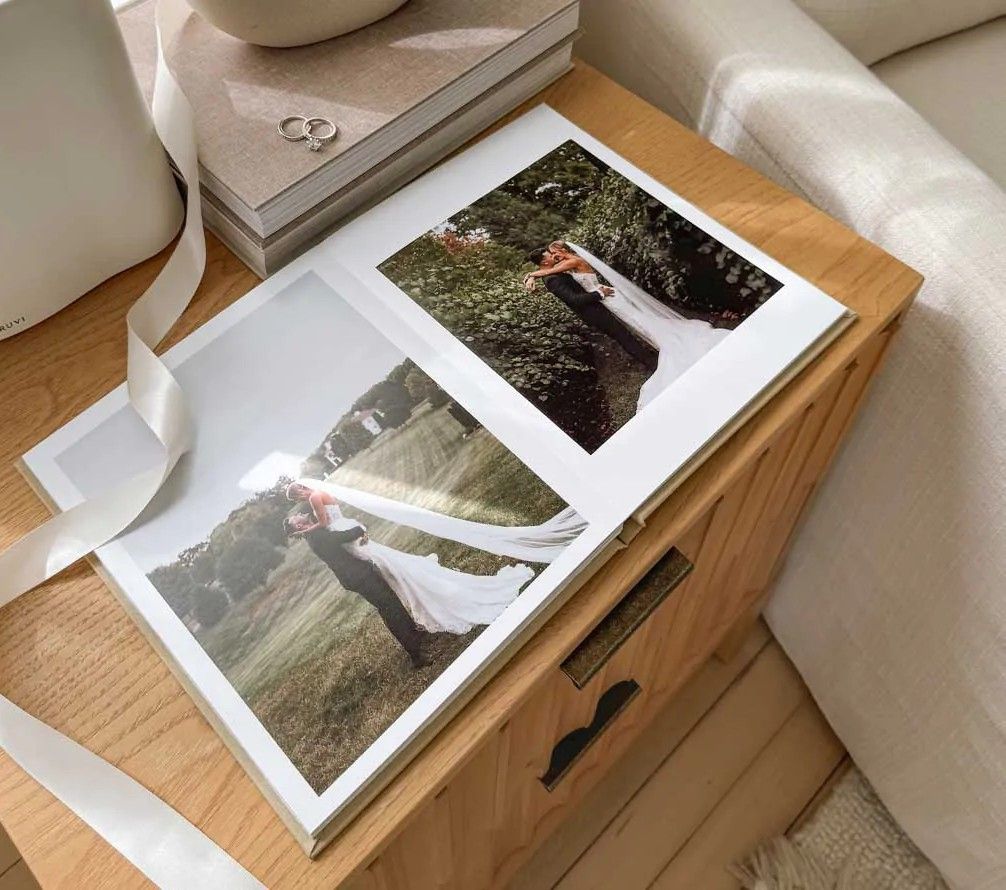 Open wedding photo album displaying bride and groom portraits on a wooden dresser with white ribbon and ring box.